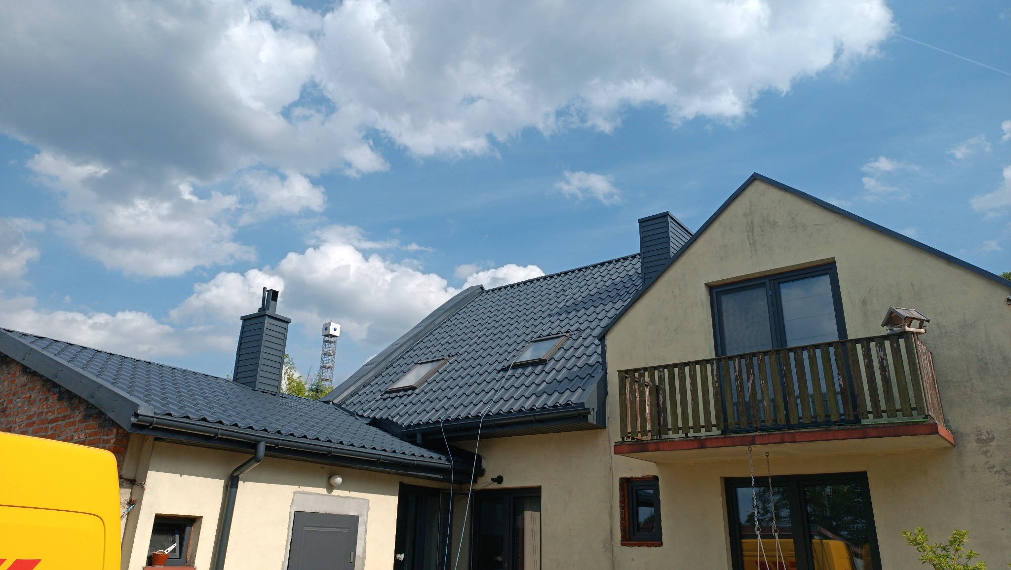 Elewacja domu z balkonem i ciemnoszarą dachówką na tle błękitnego nieba z chmurami. Widoczny fragment żółtego samochodu dostawczego.