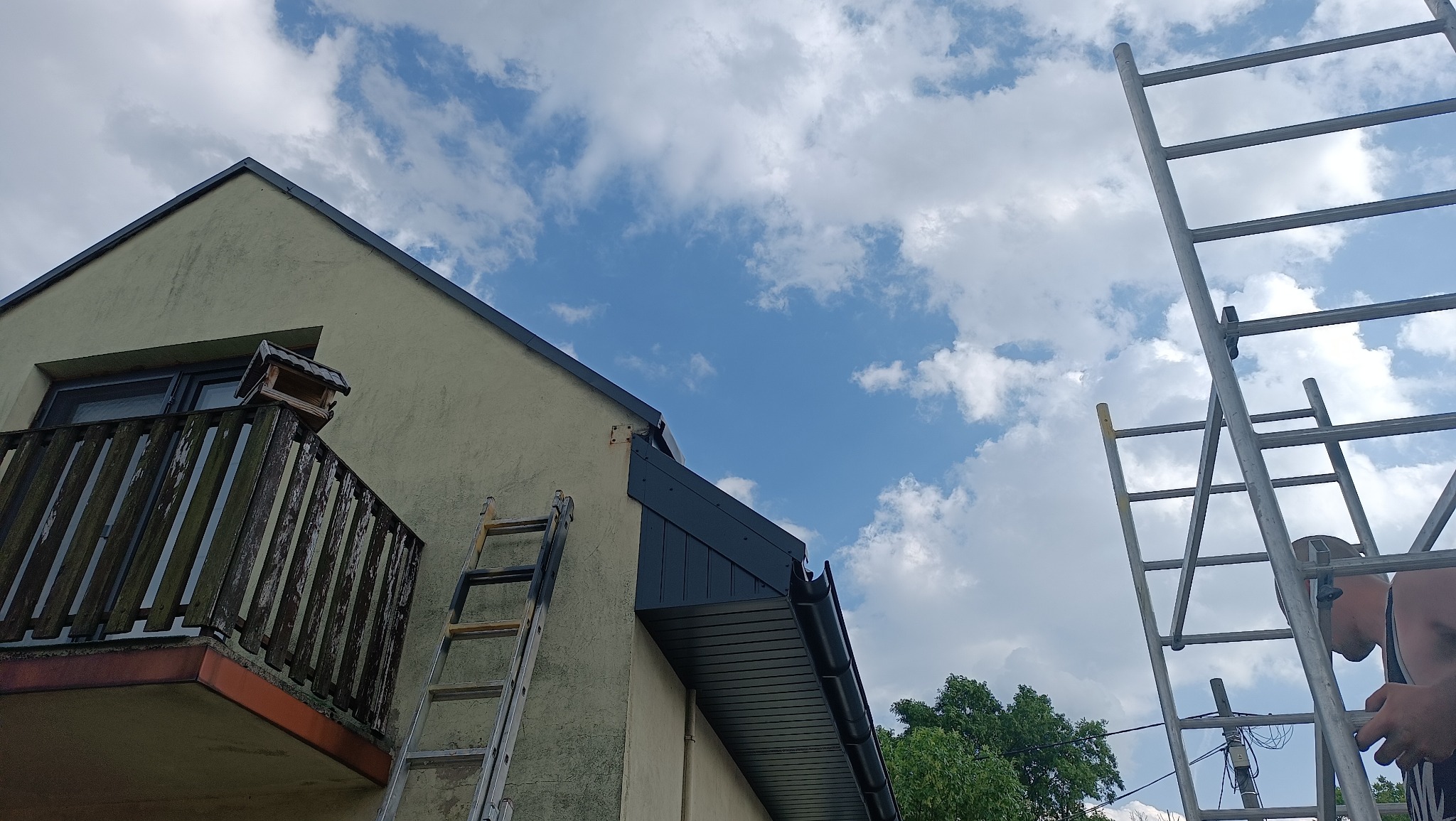 Fragment elewacji domu z zamontowanym, ciemnym podsufitkiem. Dwie drabiny opierają się o ścianę. Na balkonie karmnik dla ptaków. Błękitne niebo z chmurami w tle.