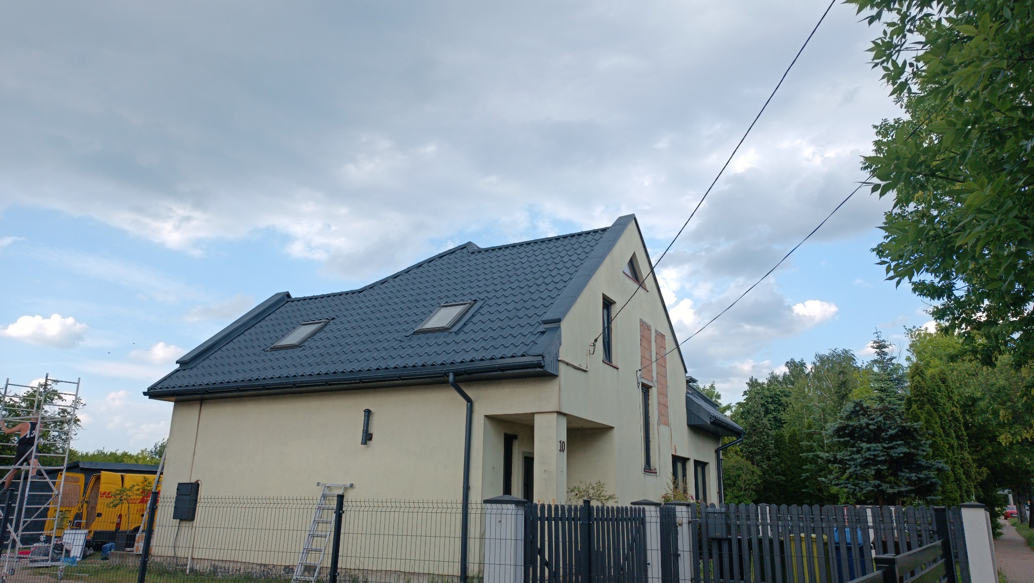 Dom jednorodzinny po renowacji z ciemnym dachem i jasną elewacją, widoczny z boku, z fragmentem rusztowania i drabiny. W tle żółty samochód dostawczy.