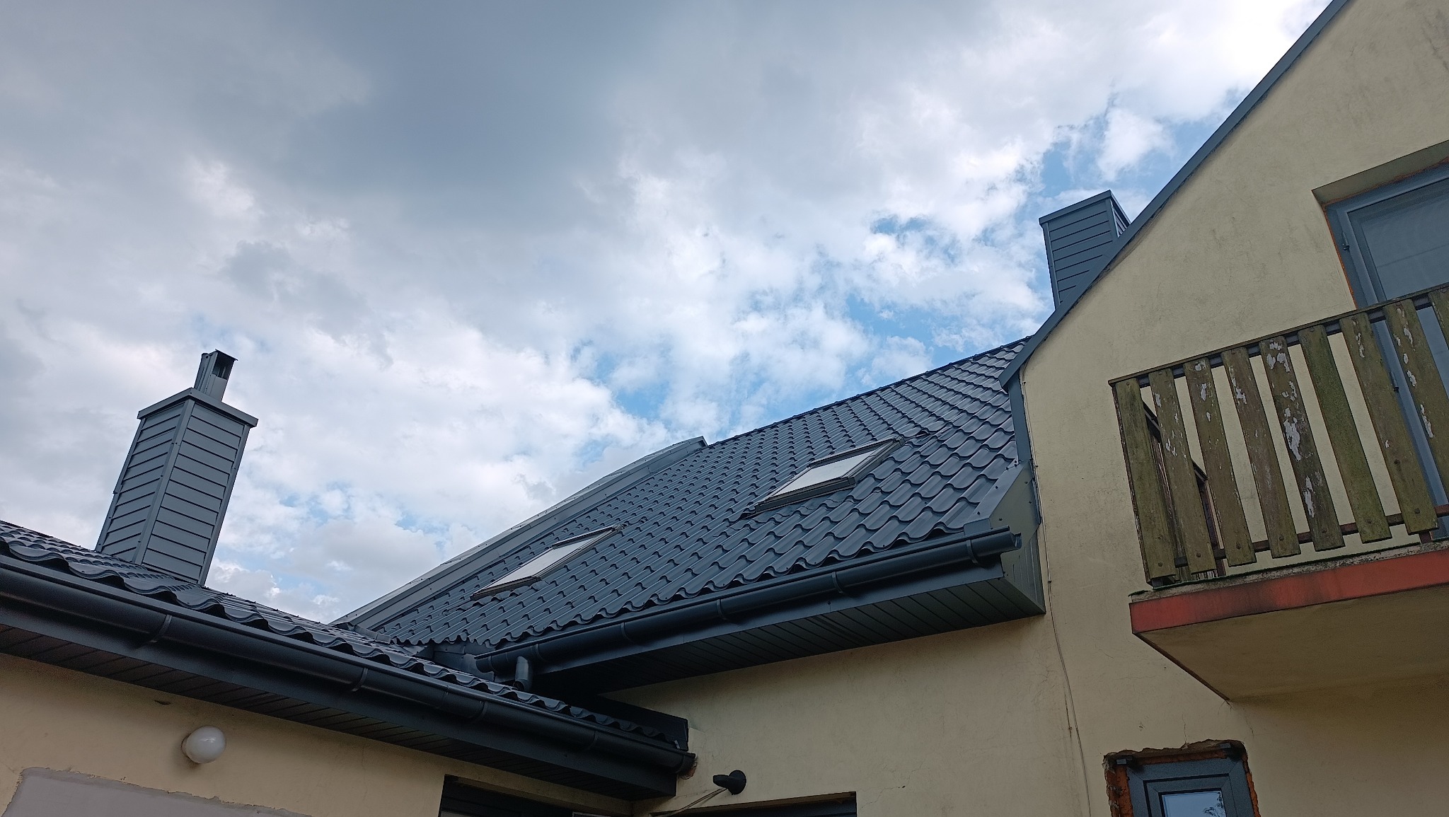 Szary dach z dachówką i kominy na tle pochmurnego nieba, widoczne okna dachowe i fragment elewacji z balkonem. Ujęcie z dołu.