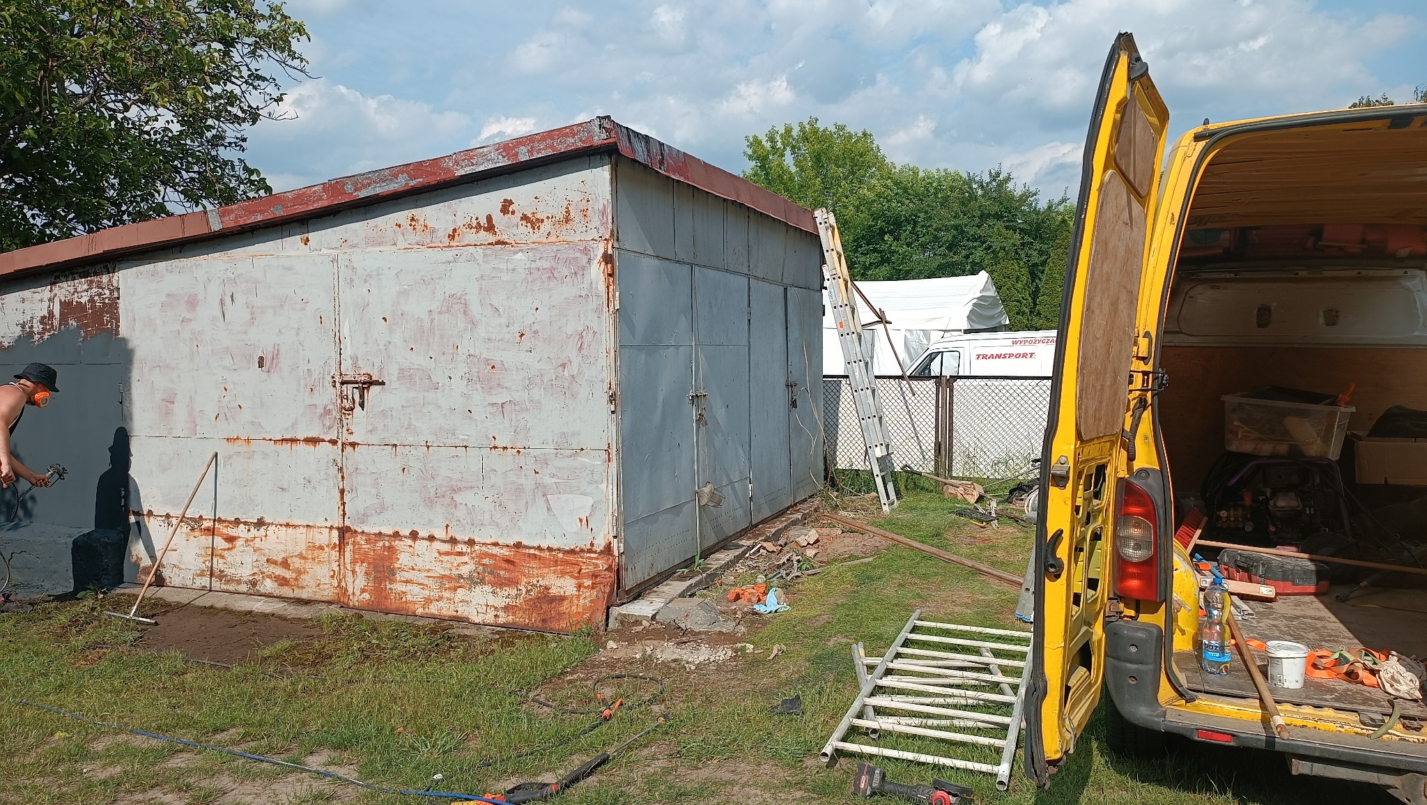 Malowanie metalowego garażu z widoczną rdzą przez osobę w masce, z otwartym żółtym vanem w tle, drabina oparta o ścianę, teren trawiasty.