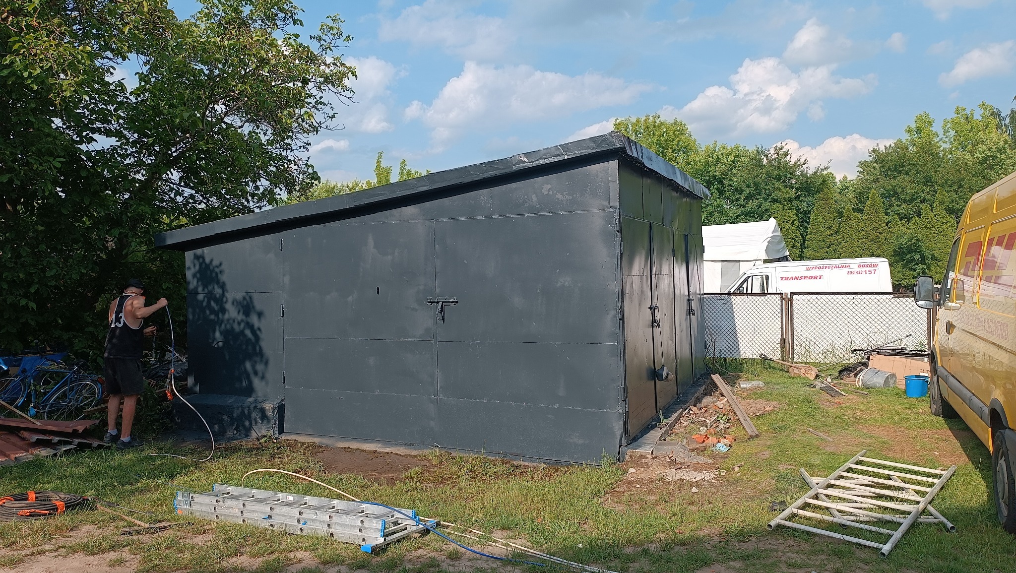 Malowanie metalowego garażu na szaro. Mężczyzna z wężem ogrodowym w tle, drabina i inne narzędzia na trawie. Widoczne ślady farby na ziemi i ścianie.