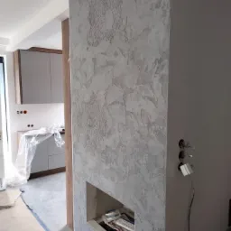 Ściana z dekoracyjną, szarą fakturą i ornamentami, w tle widoczna kuchnia w trakcie prac wykończeniowych, zabezpieczona folią malarską.