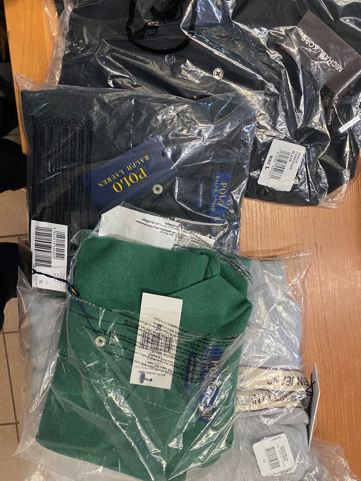 Paczki z nową odzieżą marek Ralph Lauren, Michael Kors i Calvin Klein, zapakowane w przezroczyste plastikowe torby, widoczne metki i rozmiary.