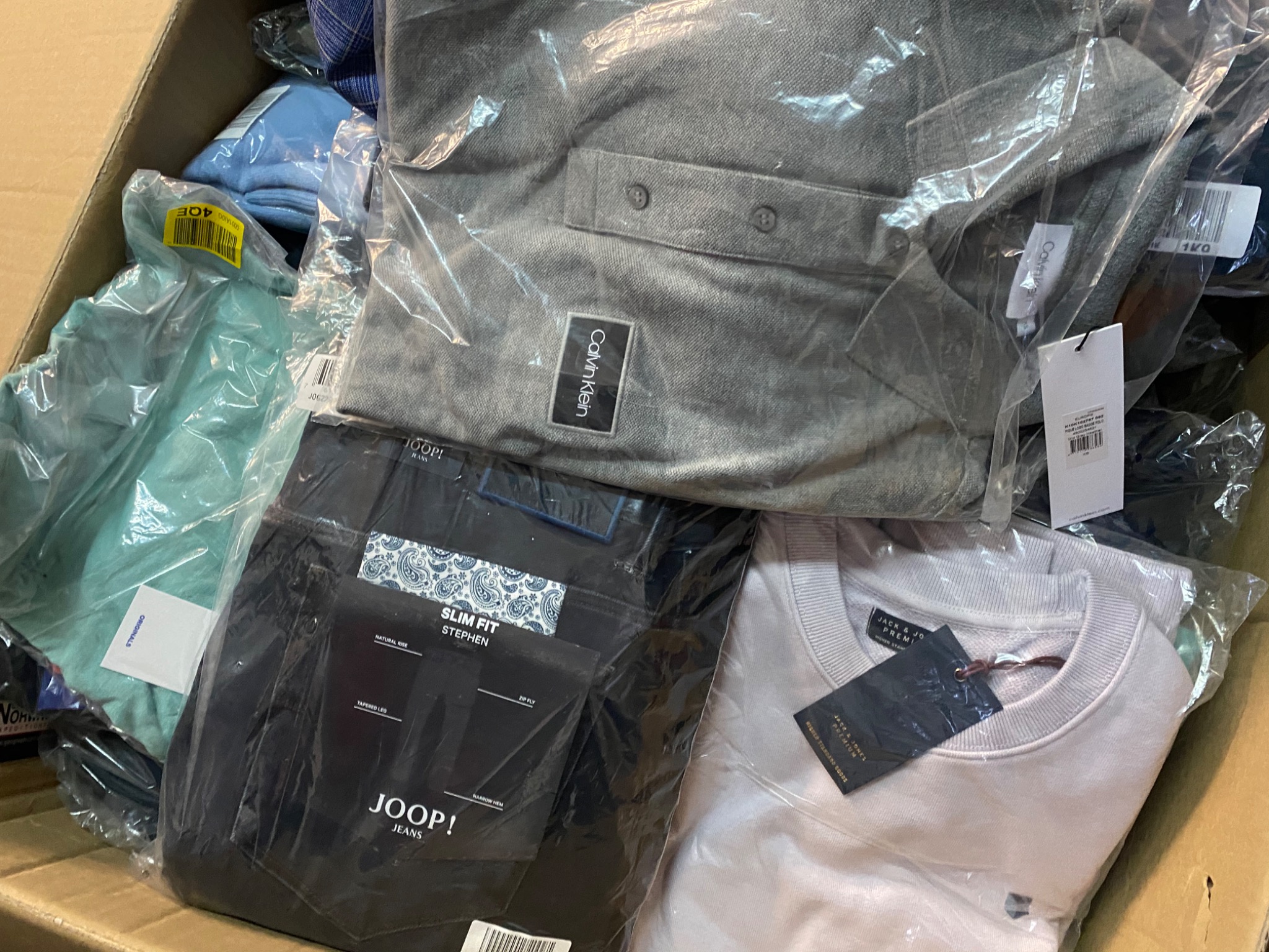 Karton wypełniony nową, markową odzieżą w foliowych opakowaniach, widoczne logo Calvin Klein, Joop! Jeans oraz Jack & Jones Premium.