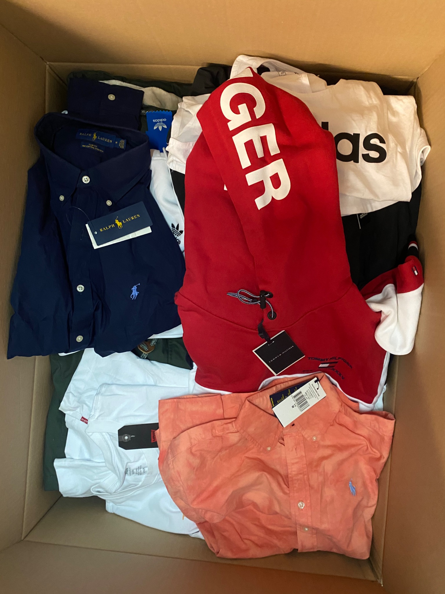 Kartonowe pudło wypełnione różnorodną odzieżą markową, w tym koszule Ralph Lauren w kolorze granatowym i łososiowym, bluza Tommy Hilfiger z kapturem oraz biała koszulka Adidas z dużym logo.