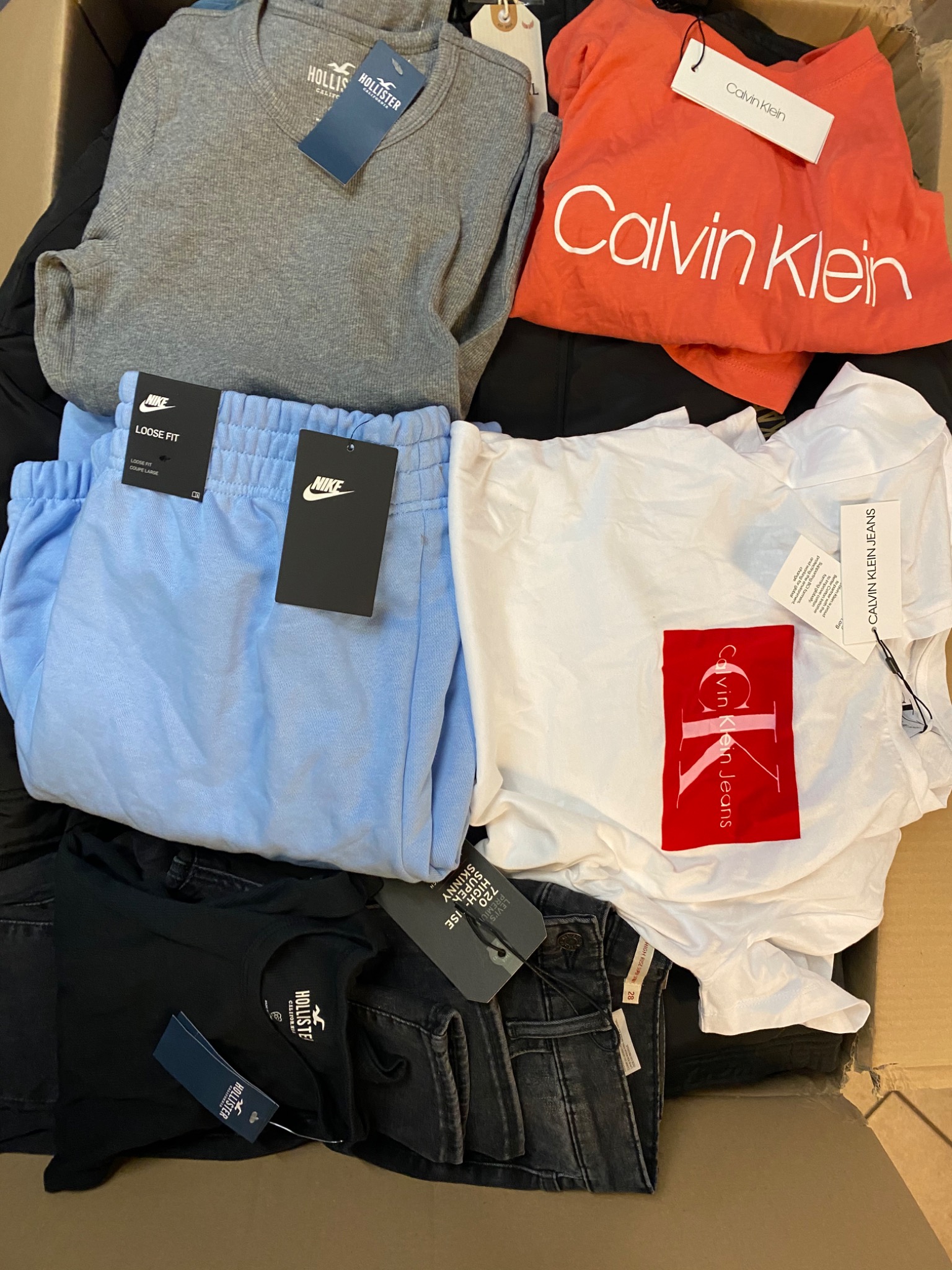 Różnorodne ubrania markowe w kartonowym pudle: szara bluza Hollister, pomarańczowa koszulka Calvin Klein, błękitne spodnie Nike, biała koszulka Calvin Klein Jeans z czerwonym logo i czarne jeansy...