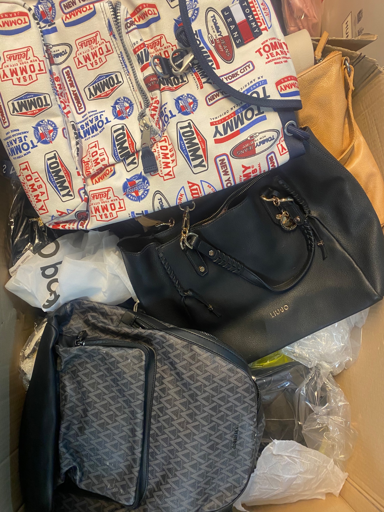 Używane torebki różnych marek i wzorów, w tym czarna torebka Liu Jo z plecionymi uchwytami, torba Valentino z geometrycznym wzorem i kurtka Tommy Jeans z nadrukami logo, ułożone w kartonowym pudle.