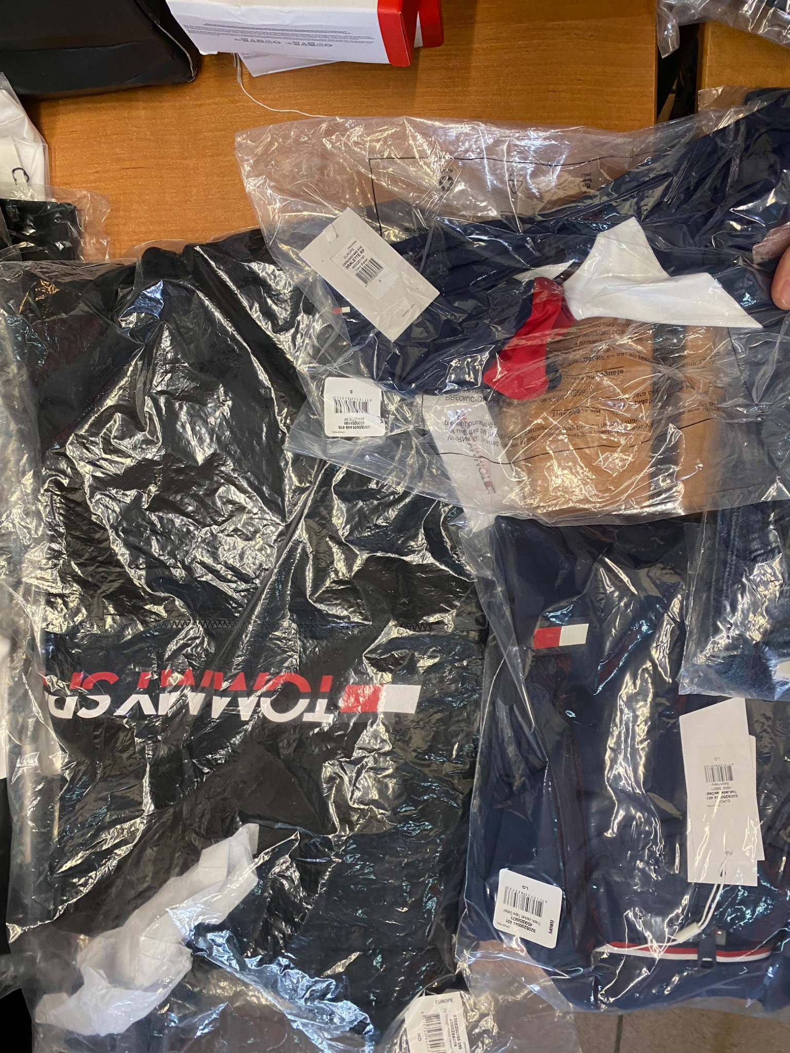 Nowa odzież marki Tommy Hilfiger zapakowana w folię, widoczne metki i rozmiary, ułożona na drewnianym blacie.