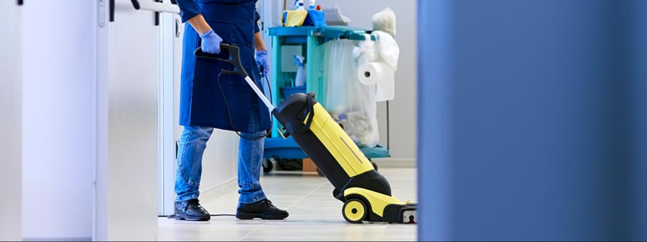 Pracownik w niebieskim uniformie i rękawicach używa żółto-czarnego odkurzacza Kärcher na jasnej podłodze w biurze; w tle wózek z akcesoriami do sprzątania.