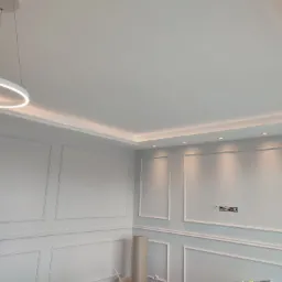 Wykończone wnętrze w stylu klasycznym z białymi ścianami ozdobionymi sztukaterią, nowoczesnym okrągłym oświetleniem sufitowym i ukrytym oświetleniem LED wzdłuż sufitu. Widoczna puszka elektryczna...