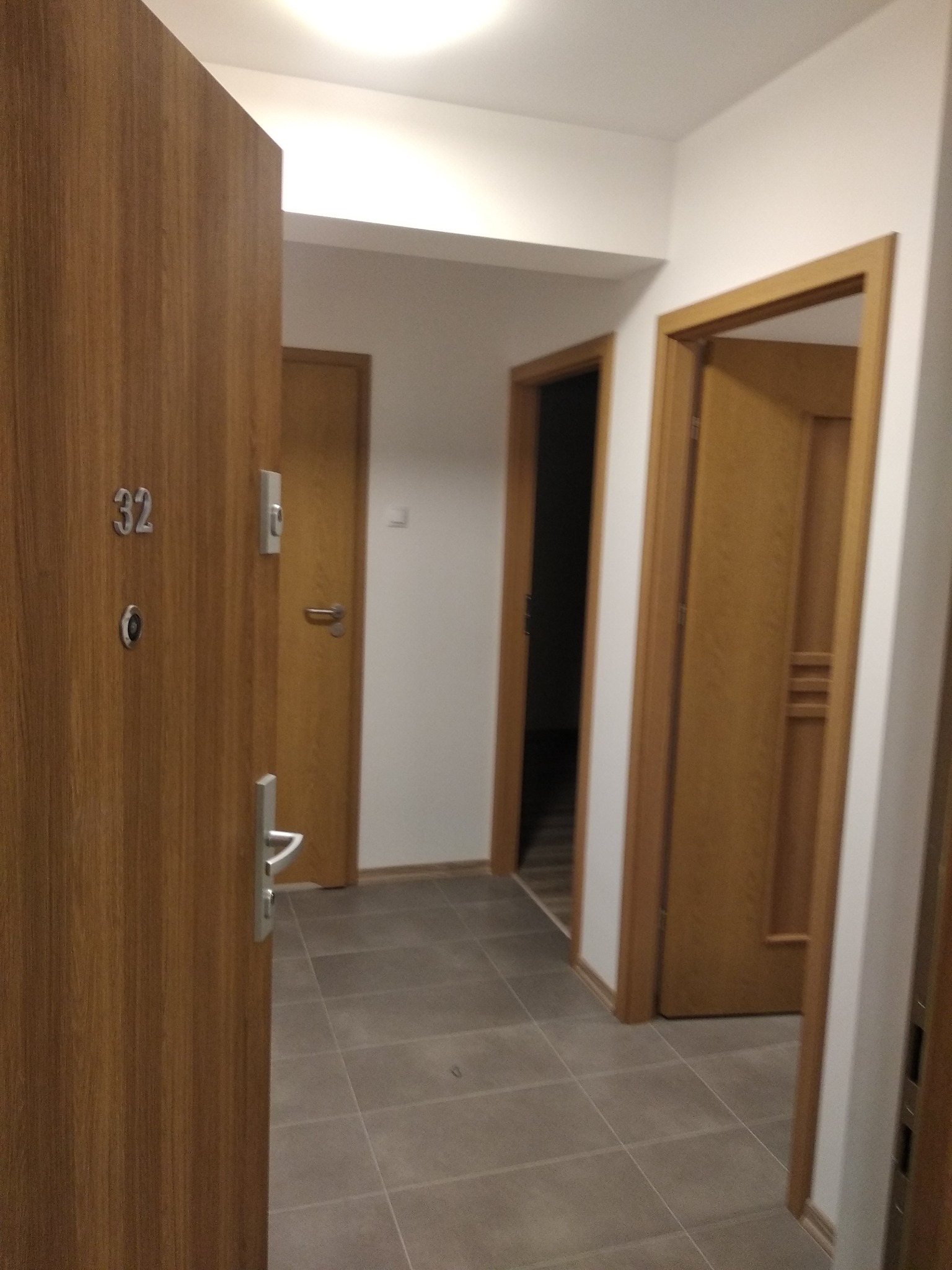 Wnętrze korytarza z trzema drewnianymi drzwiami, szara płytka podłogowa, widok zza otwartych drzwi wejściowych z numerem '32'.