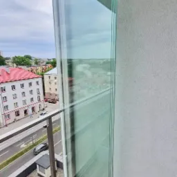 Zastosowanie przesuwnych paneli szklanych w zabudowie balkonu, widok z góry na drewnianą podłogę balkonu i fragment zabudowy miejskiej w tle.