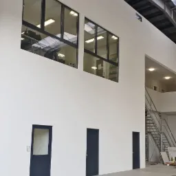 Wnętrze hali przemysłowej z trzema ciemnymi drzwiami i dwoma oknami aluminiowymi umieszczonymi wysoko na białej ścianie, widoczne schody metalowe w głębi.