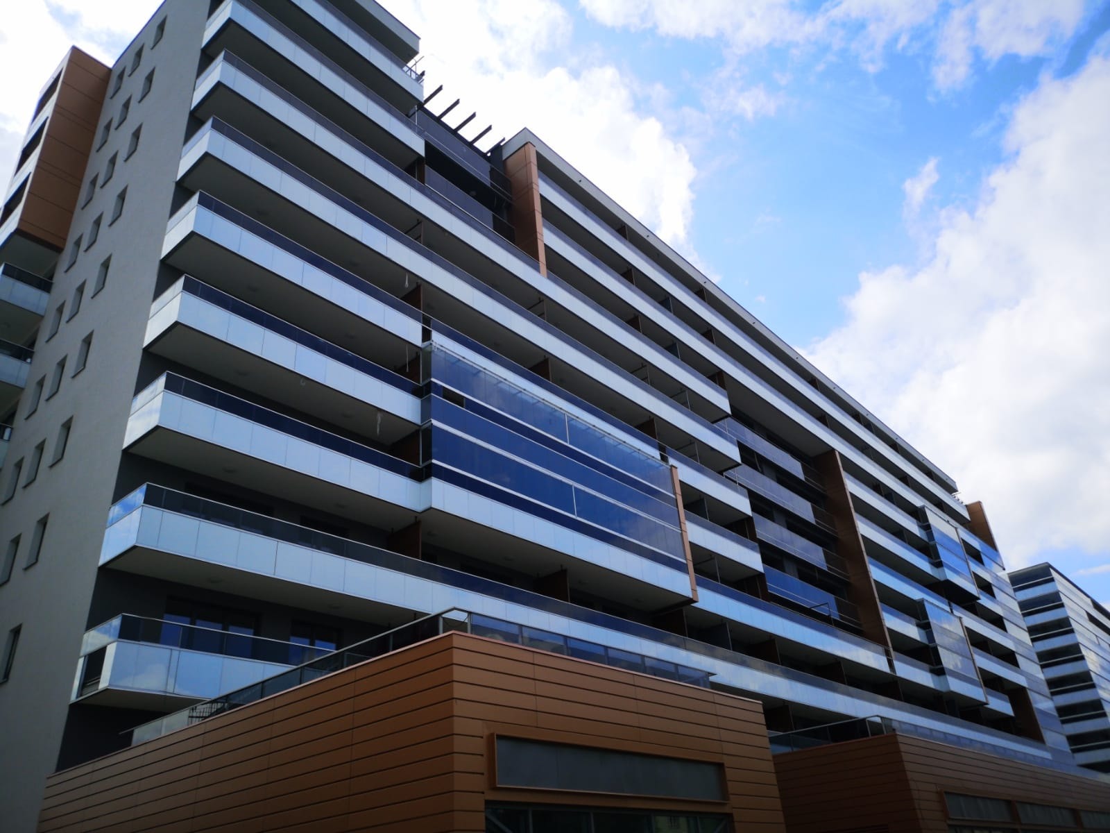 Nowoczesny budynek mieszkalny z balkonami z balustradami szklanymi, widok z dołu na błękitne niebo z chmurami.