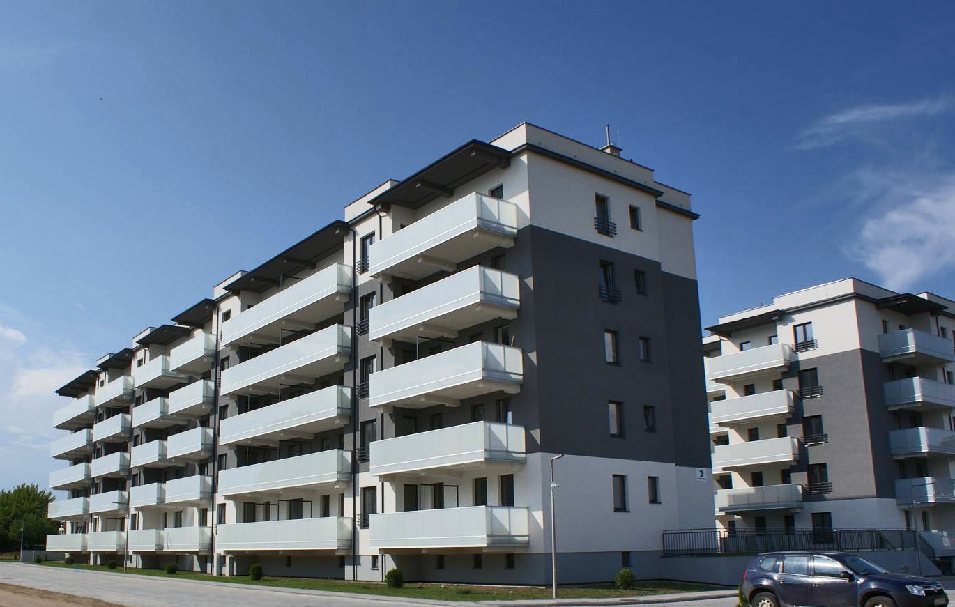 Nowoczesny budynek wielorodzinny w Olecku z balkonami zabezpieczonymi mlecznobiałymi balustradami, widok z poziomu ulicy w słoneczny dzień.