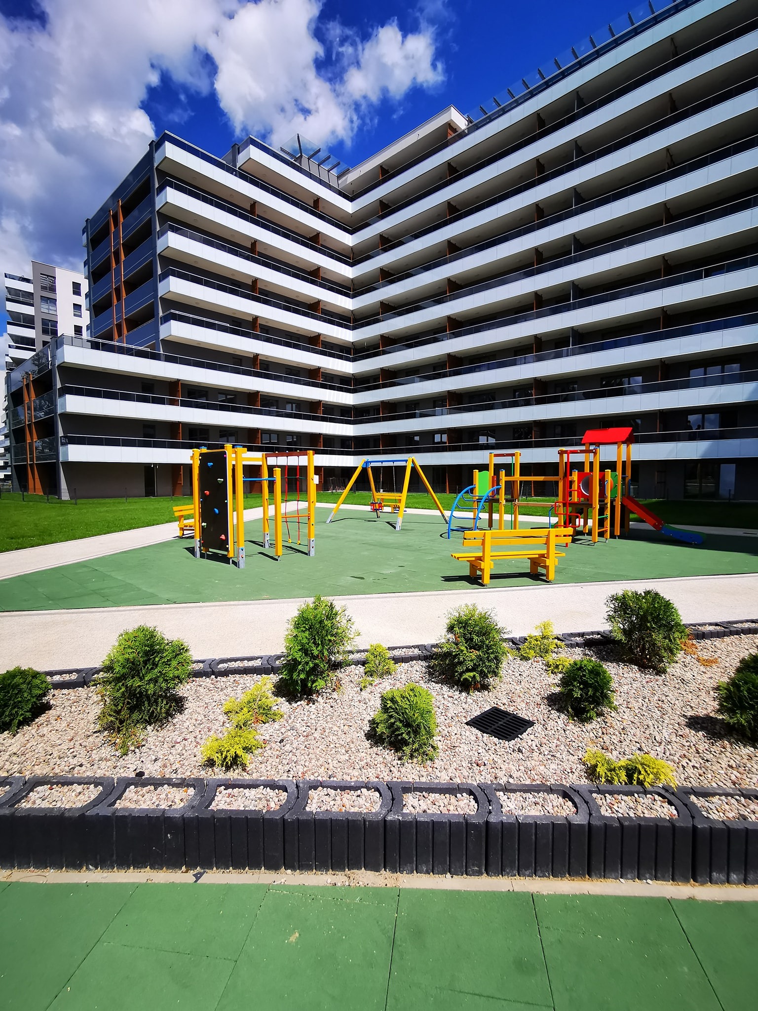 Nowoczesny plac zabaw z kolorowymi elementami na tle wielopiętrowego budynku mieszkalnego z balkonami, otoczony zielenią i ozdobnymi krzewami