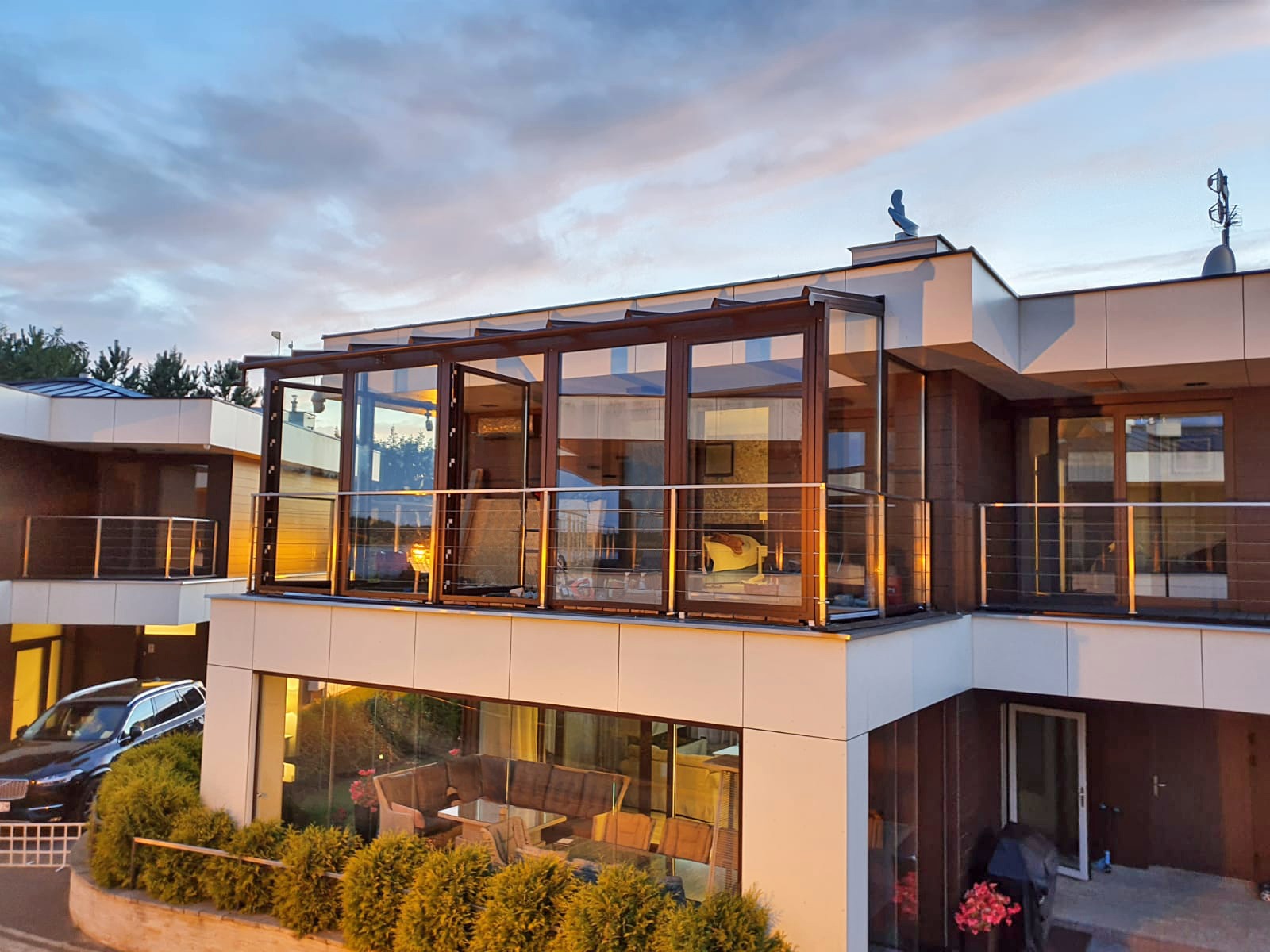 Zadaszony taras na piętrze z przesuwnymi szklanymi ścianami i balustradą z cienkich, poziomych prętów, widok z dołu na tle zachodzącego słońca.