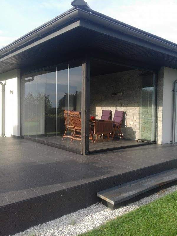 Zewnętrzna szklana zabudowa tarasu z przesuwnymi panelami, czarne ramy, ciemna podłoga z płytek, widoczne meble ogrodowe wewnątrz.