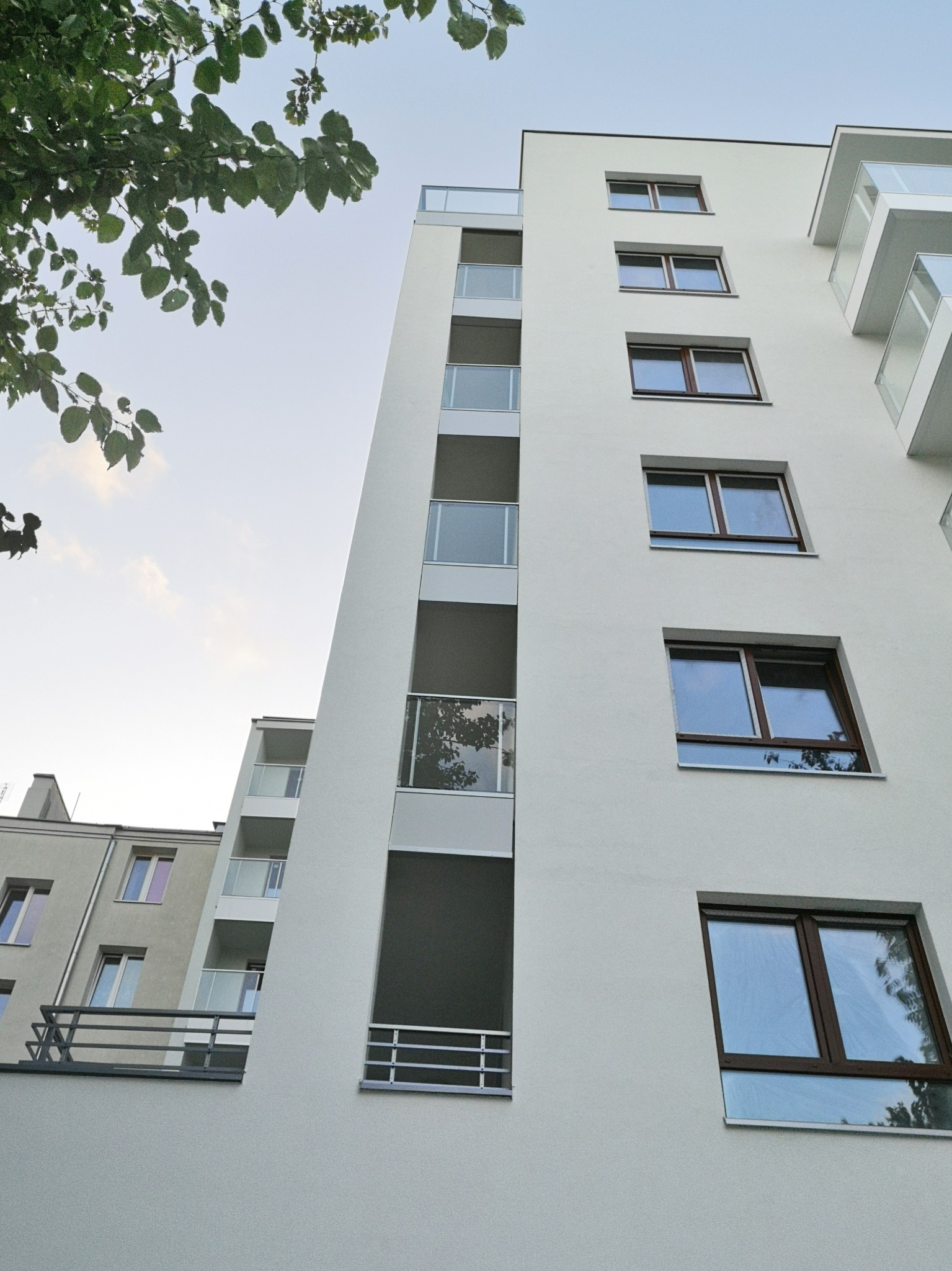 Biały budynek wielorodzinny z balkonami z przezroczystymi balustradami i brązowymi ramami okiennymi, widziany z dołu, z fragmentem drzewa w lewym górnym rogu.