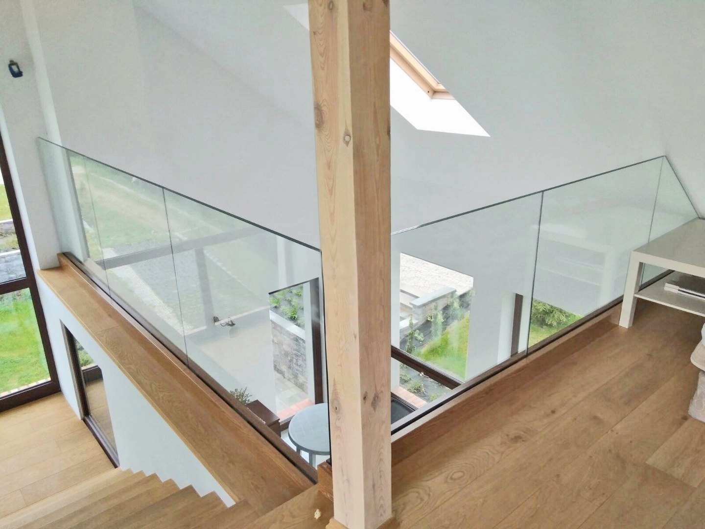 Szklana balustrada na antresoli z drewnianymi elementami, widok z góry na schody i otoczenie domu przez duże okna.