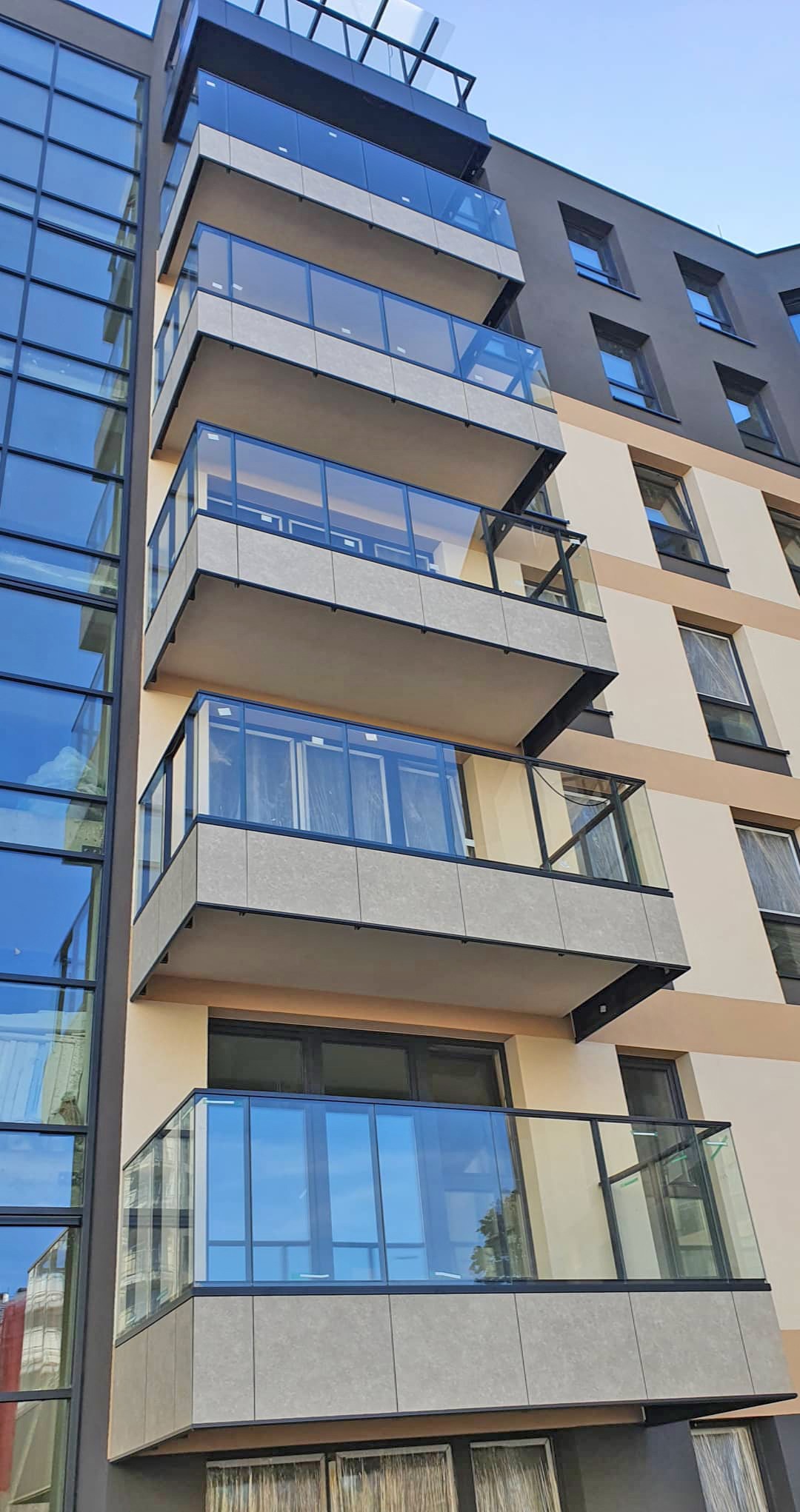 Nowoczesny budynek z balkonami z balustradami szklanymi, widok z dołu na fasadę z elementami w kolorze beżowym i szarym.