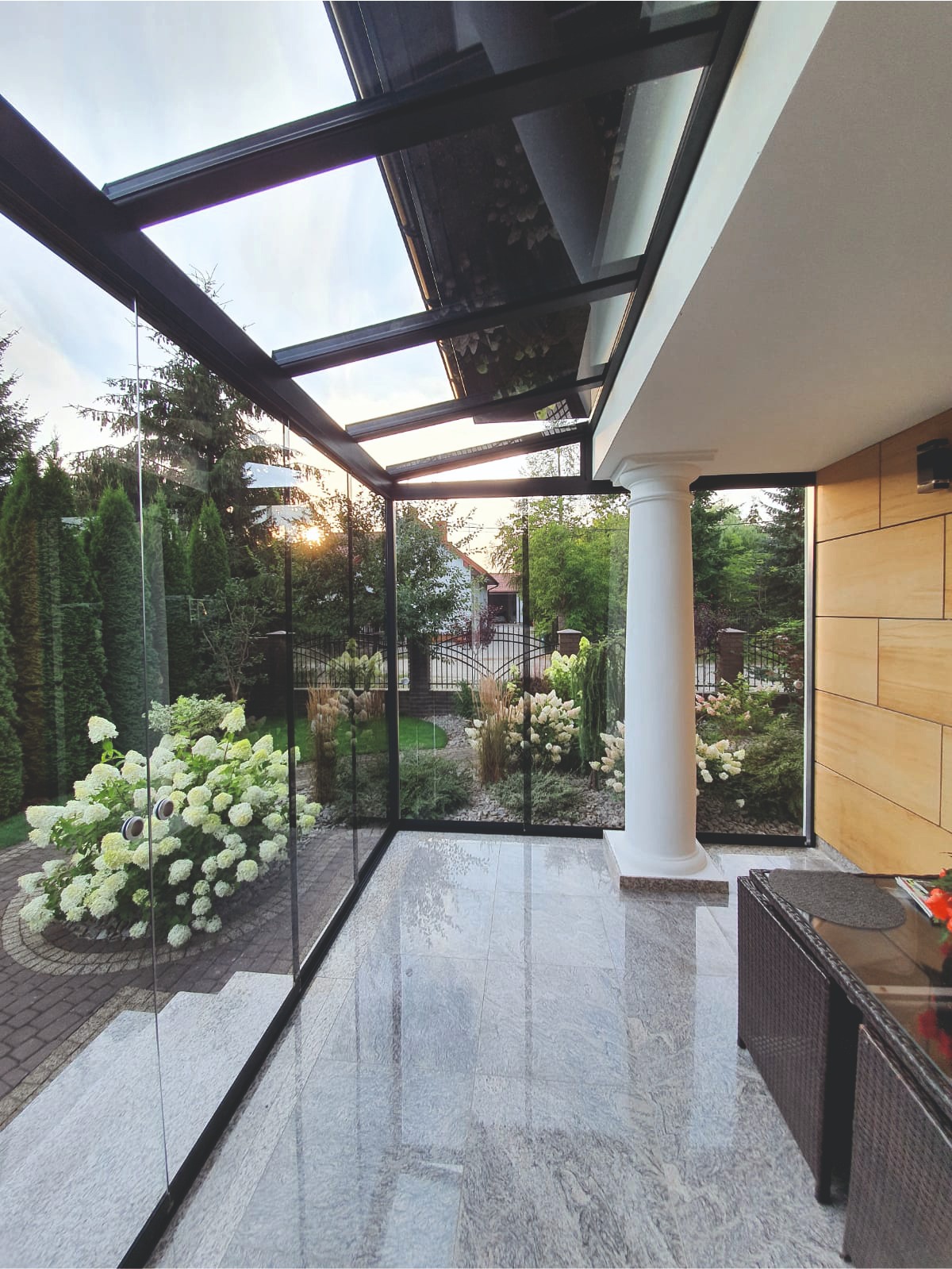 Zadaszony taras z przeszklonymi ścianami i dachem, widok na ogród z kwitnącymi hortensjami, marmurowa posadzka odbija światło zachodzącego słońca.