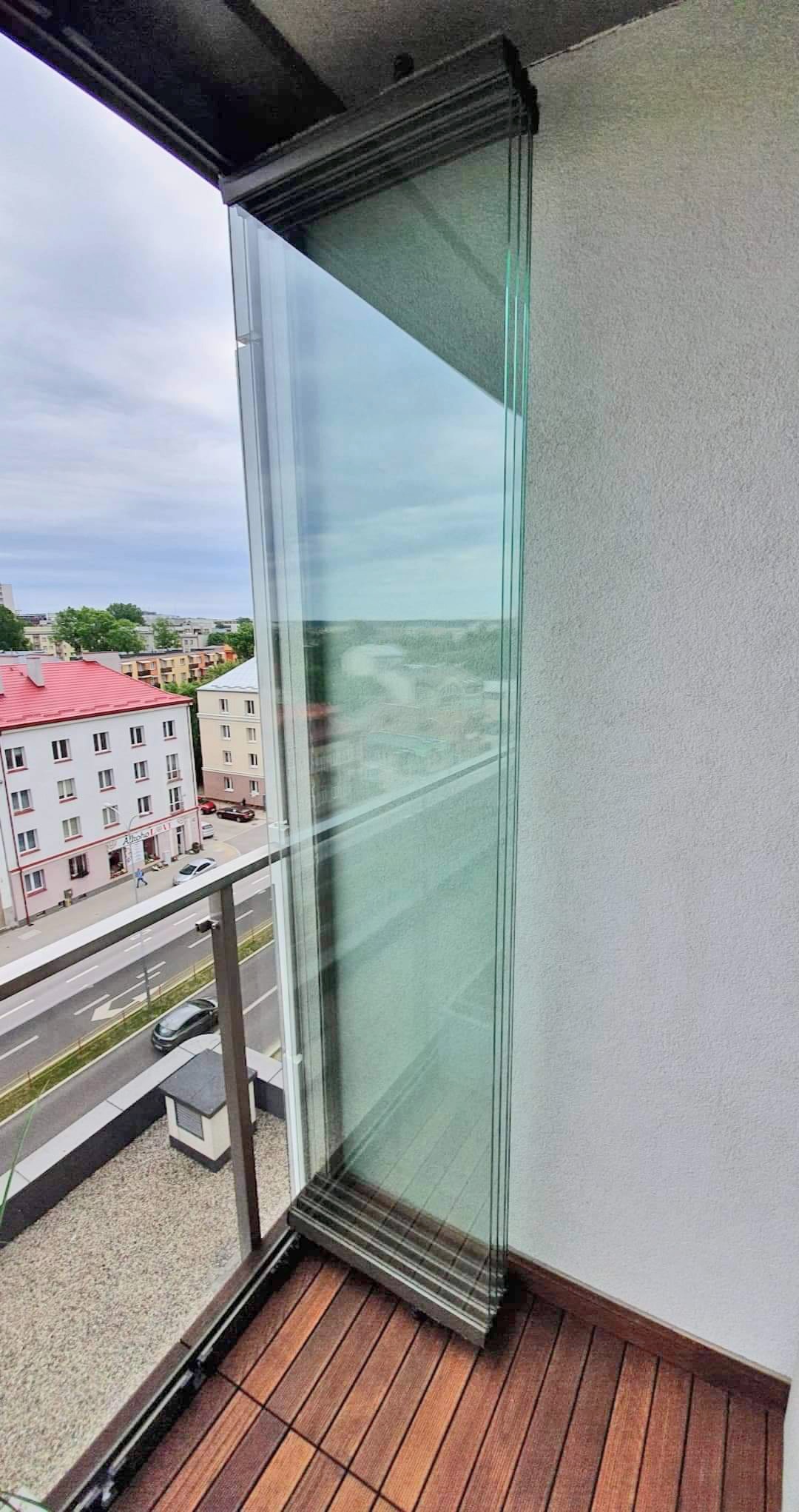 Zastosowanie przesuwnych paneli szklanych w zabudowie balkonu, widok z góry na drewnianą podłogę balkonu i fragment zabudowy miejskiej w tle.