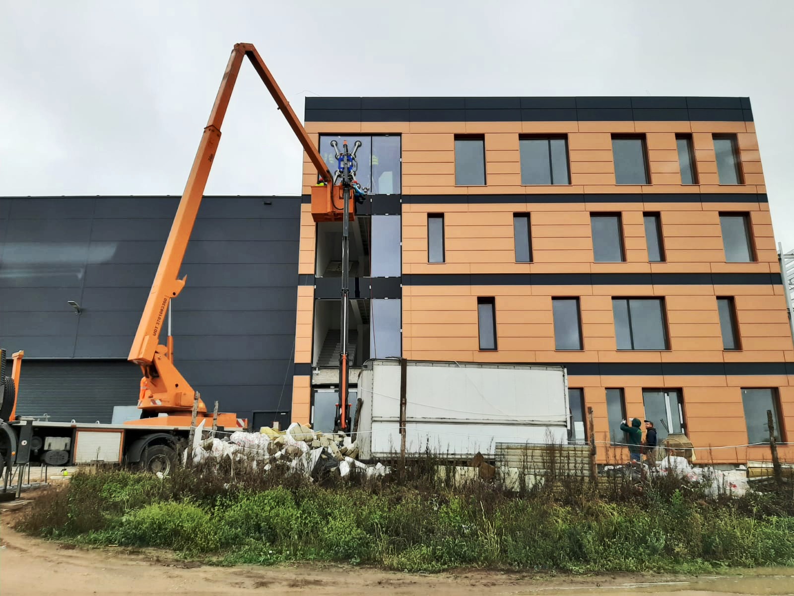 Montaż okien na elewacji budynku biurowego za pomocą pomarańczowego podnośnika koszowego; widoczni pracownicy w koszu i na ziemi.