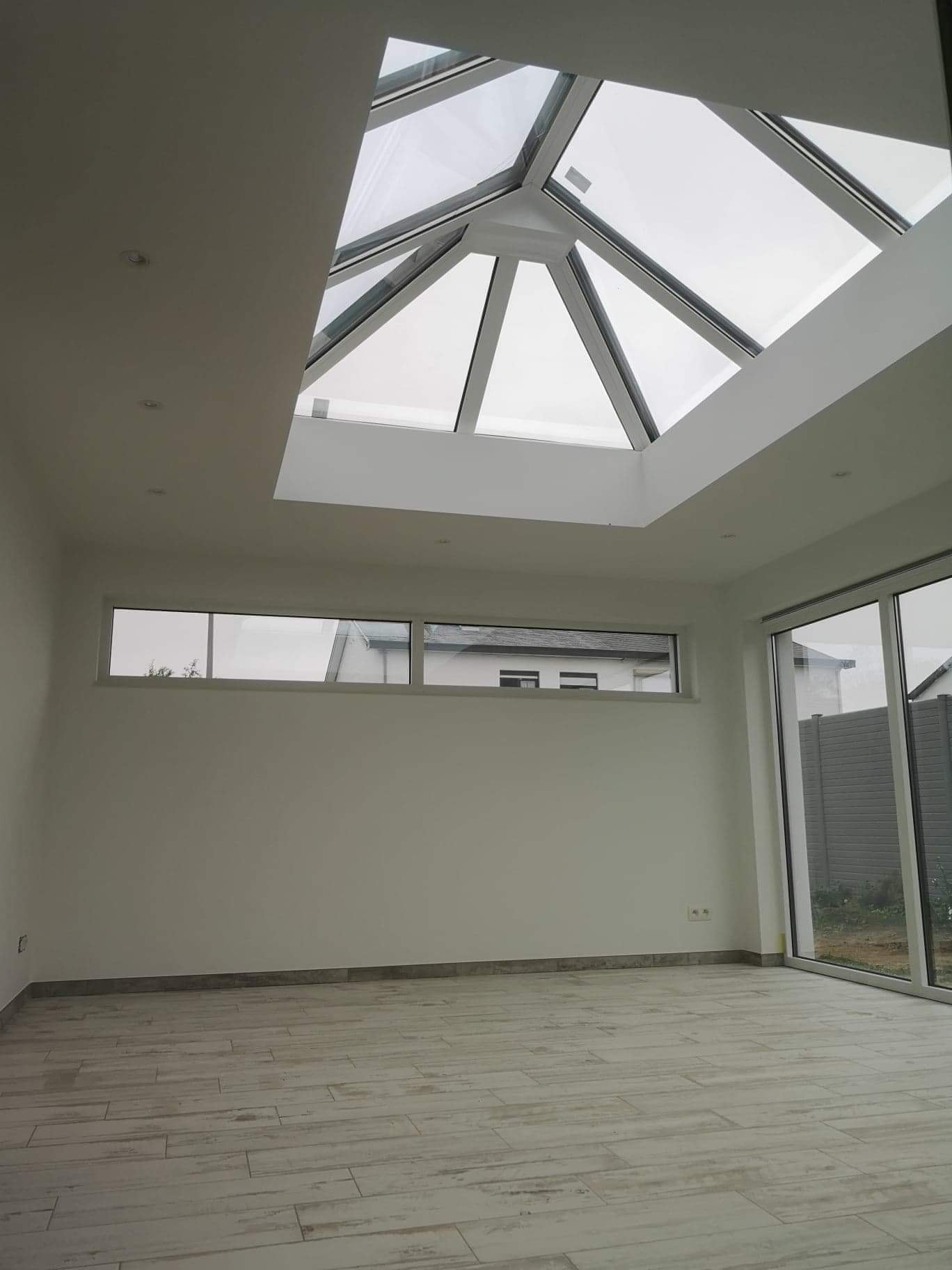 Nowoczesne wnętrze domu z dużym oknem dachowym o geometrycznym kształcie i aluminiową ramą, widoczne okno poziome i przesuwne drzwi tarasowe z widokiem na ogród.