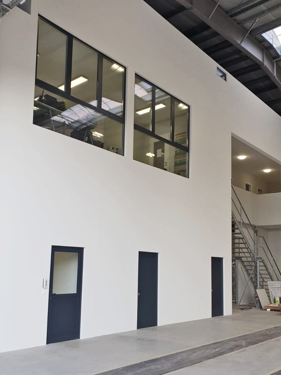 Wnętrze hali przemysłowej z trzema ciemnymi drzwiami i dwoma oknami aluminiowymi umieszczonymi wysoko na białej ścianie, widoczne schody metalowe w głębi.