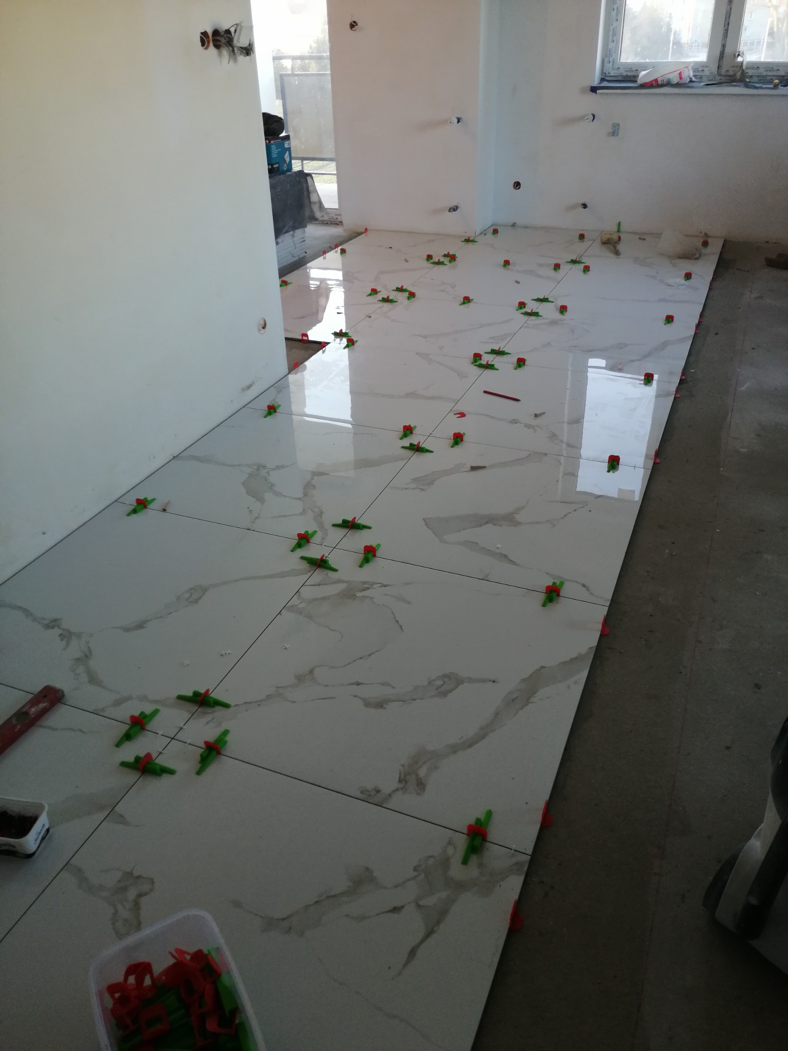 Białe płytki imitujące marmur ułożone na podłodze w trakcie prac wykończeniowych, widoczne systemy poziomowania płytek w kolorze zielono-czerwonym, ślady po zaprawie oraz niedokończone ściany.
