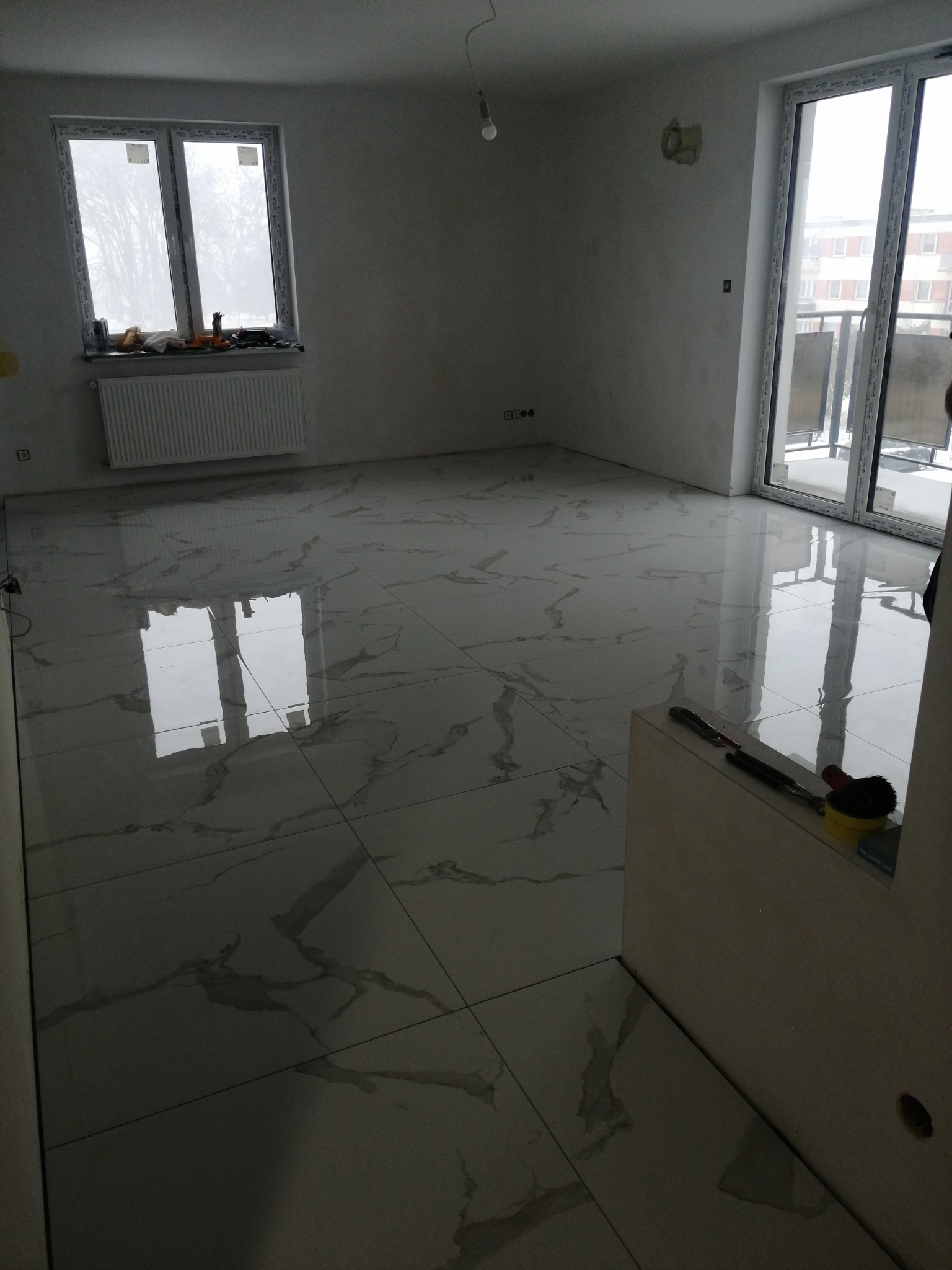 Wykończone wnętrze z błyszczącą, marmurkową podłogą z płytek, widoczne narzędzia glazurnicze przy ścianie, okno i drzwi balkonowe w tle.