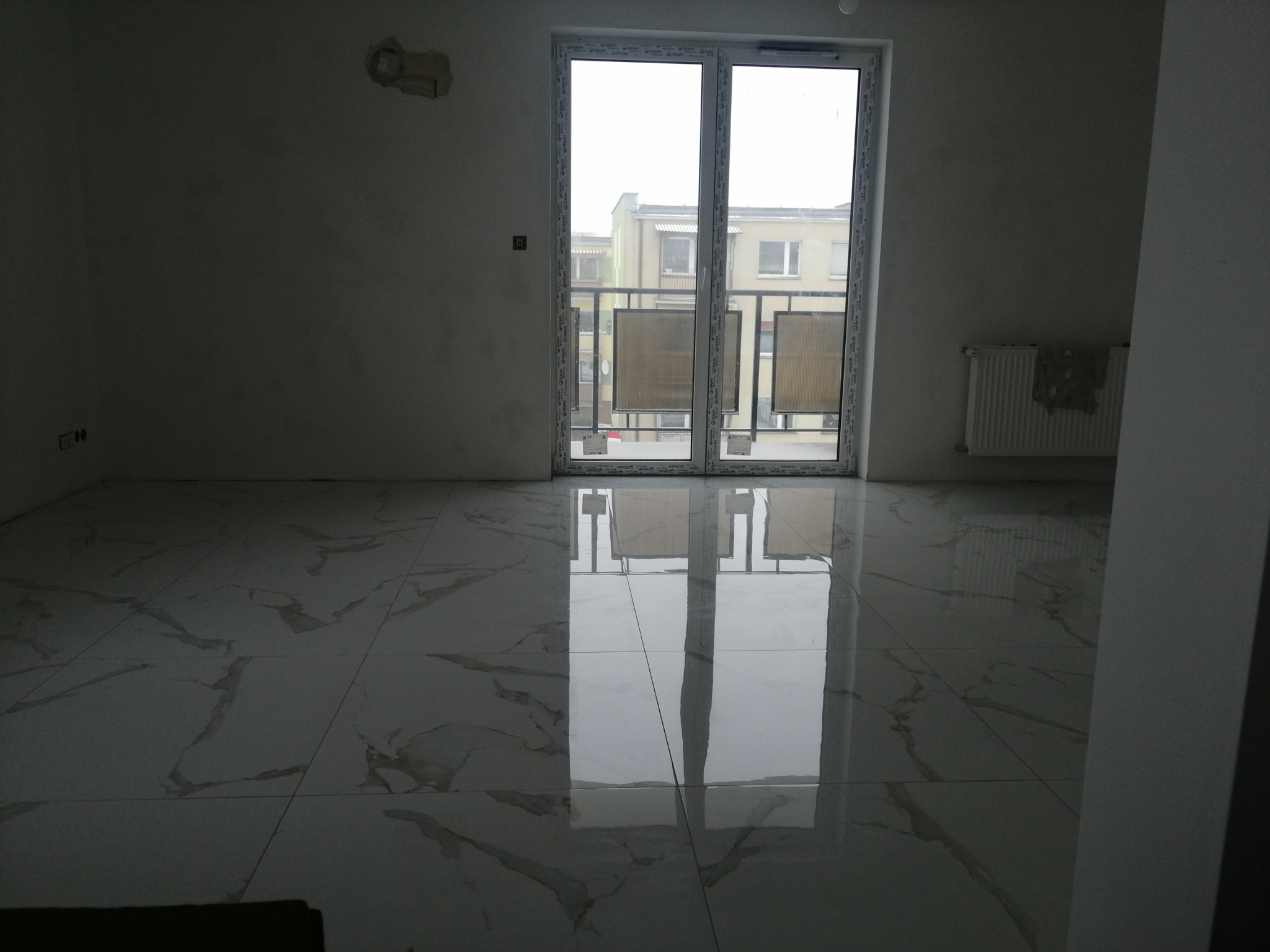 Wykończone wnętrze z błyszczącą, marmurkową podłogą w dużym formacie, odbijającą światło z okna balkonowego, ściany przygotowane do malowania, widoczny grzejnik.