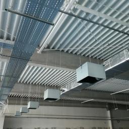 Vacuum Sp. z o.o. - Montaż systemu wentylacji w hali przemysłowej: widok na sufit z podwieszonymi kanałami wentylacyjnymi, pracownicy w odblaskowych kamizelkach nadzorują instalację.