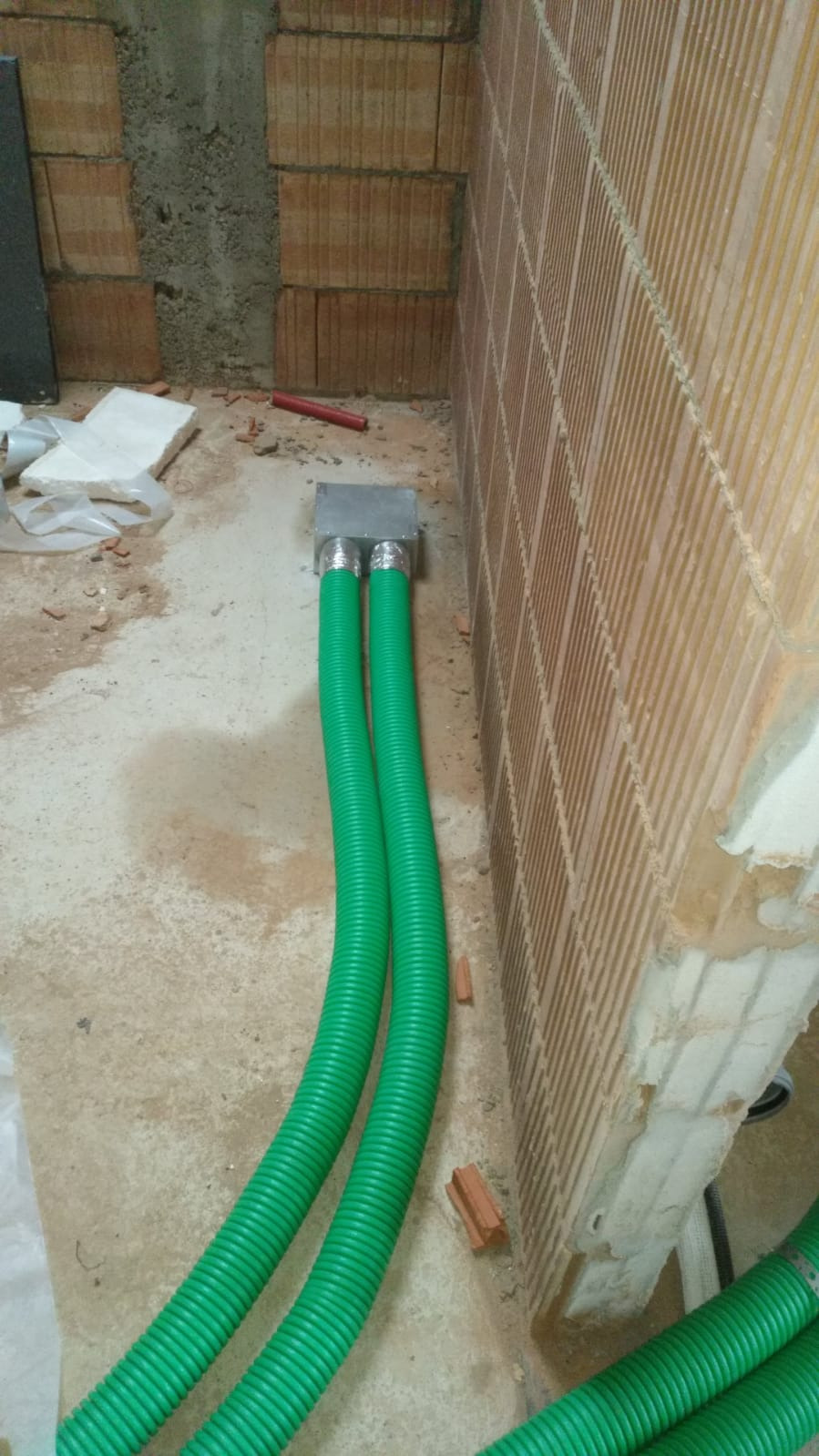 Dwa zielone, karbowane przewody wentylacyjne podłączone do metalowej skrzynki w surowym wnętrzu, ściany z cegły dziurawki, widoczne ślady budowy.