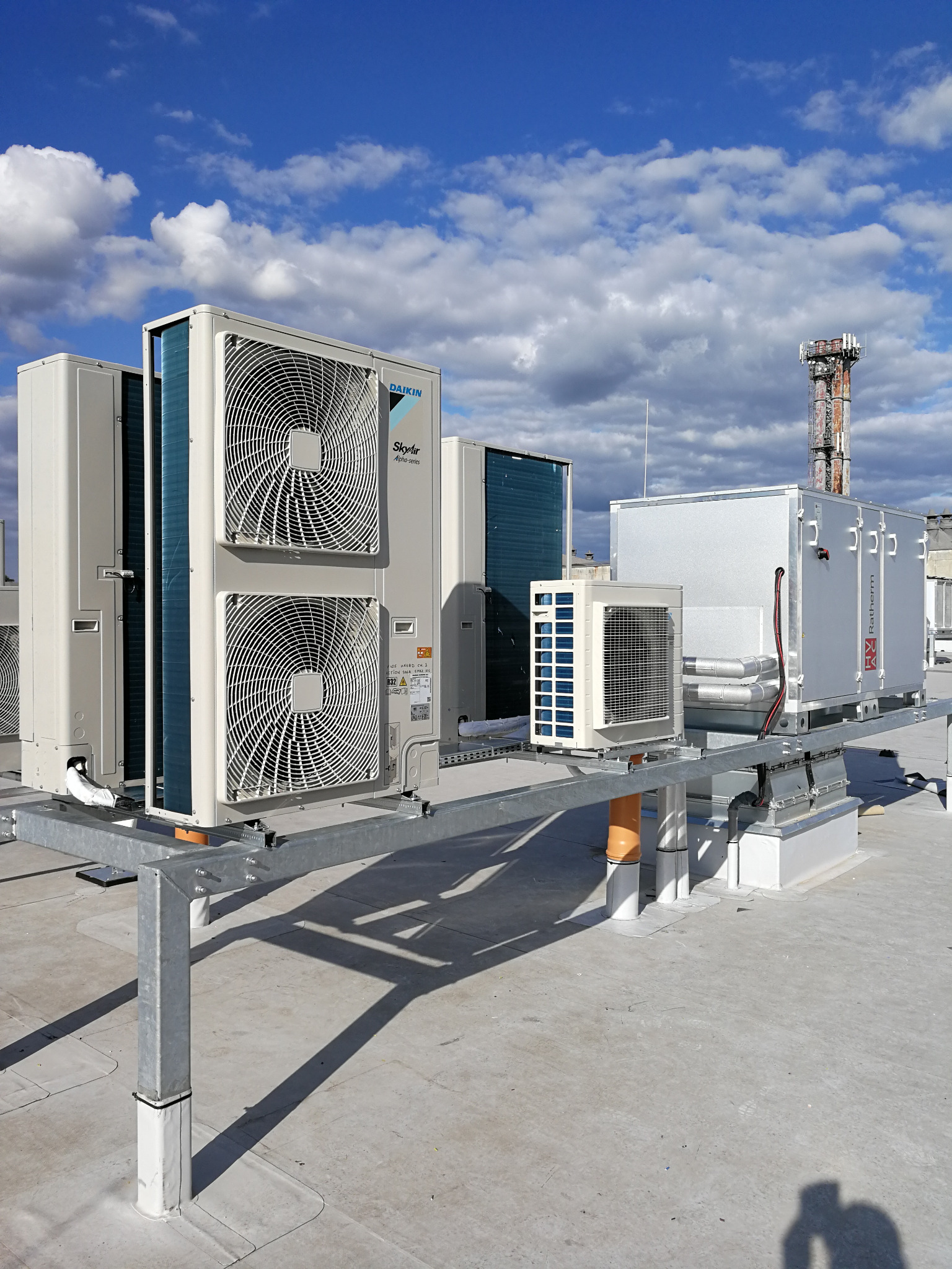 Instalacja klimatyzacji i wentylacji na dachu budynku z widocznym kominem w tle, urządzenia zamontowane na metalowej konstrukcji wsporczej.