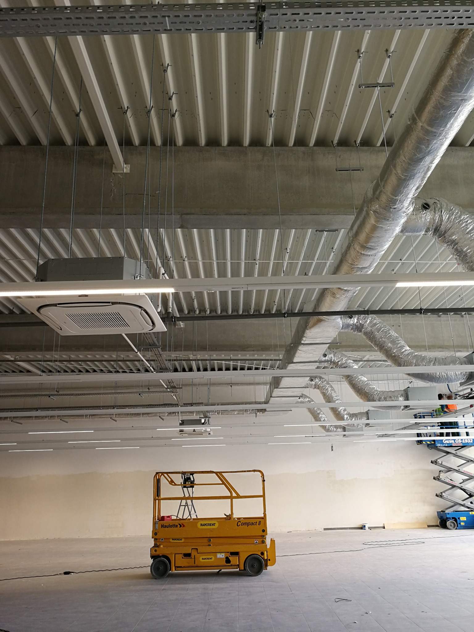 Wnętrze hali produkcyjnej z zamontowaną instalacją wentylacyjną i klimatyzacyjną, widoczne kanały wentylacyjne, klimakonwektor sufitowy oraz podnośniki nożycowe.