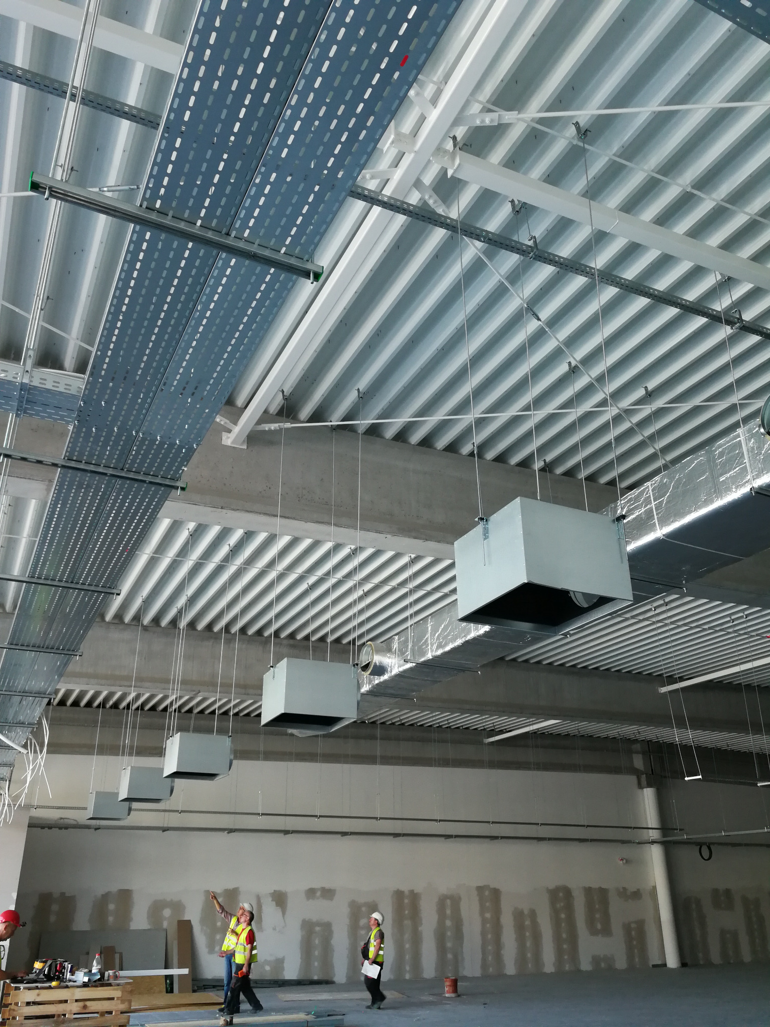 Montaż systemu wentylacji w hali przemysłowej: widok na sufit z podwieszonymi kanałami wentylacyjnymi, pracownicy w odblaskowych kamizelkach nadzorują instalację.