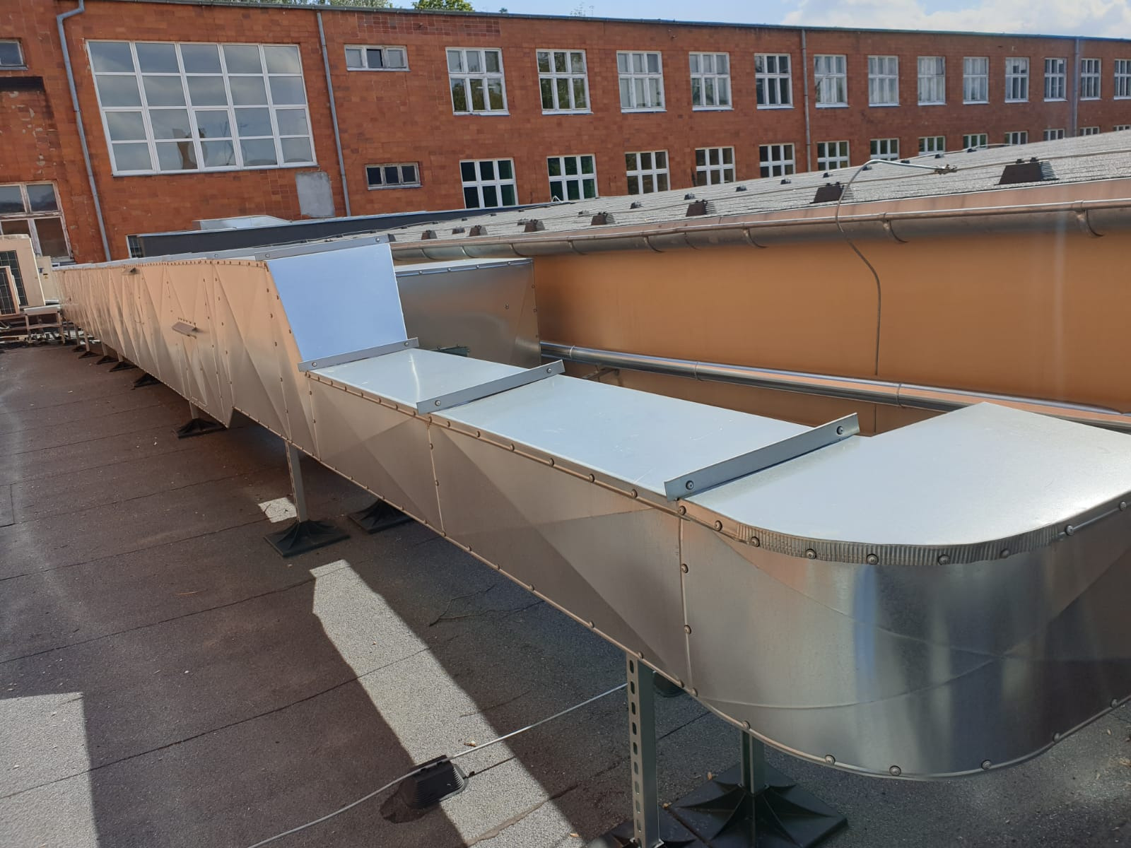 Długi, srebrny kanał wentylacyjny na dachu budynku, wsparty na metalowych stelażach z czarnymi podstawami, z ceglanym budynkiem w tle.