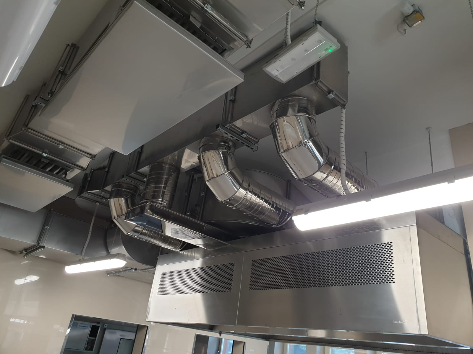 Widok z dołu na zainstalowany system wentylacyjny z metalowymi rurami i kratkami w pomieszczeniu komercyjnym, oświetlonym lampami jarzeniowymi.