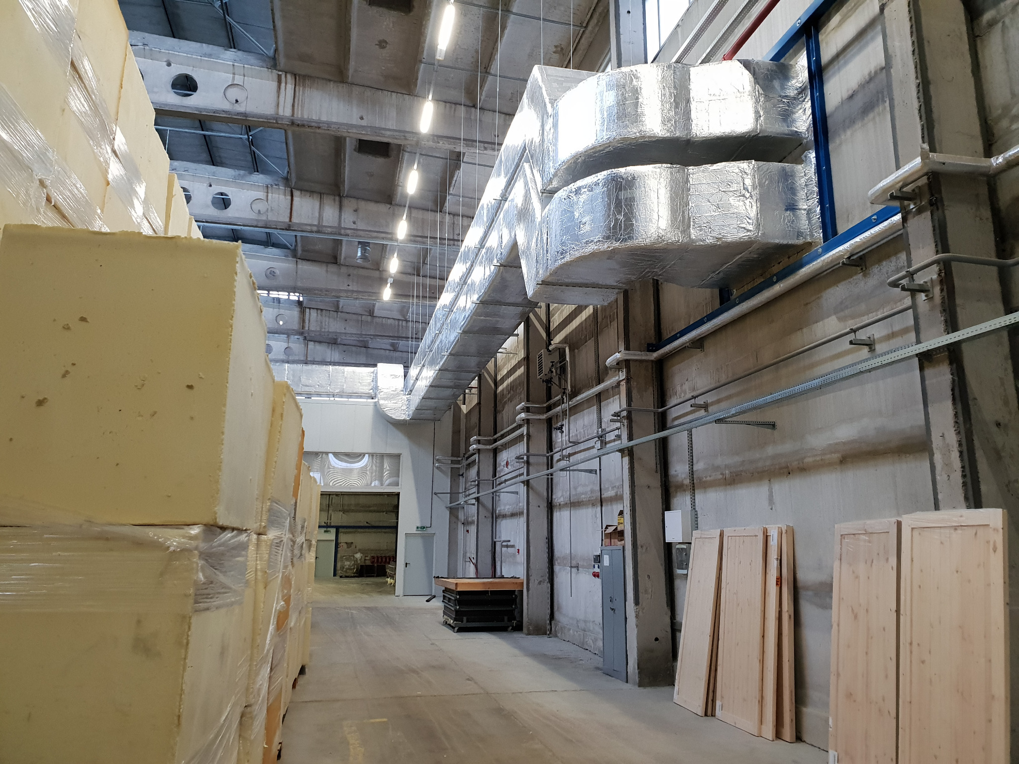 Wnętrze hali przemysłowej z widocznymi kanałami wentylacyjnymi owiniętymi folią aluminiową, ułożone bloki styropianu i elementy konstrukcyjne w tle.