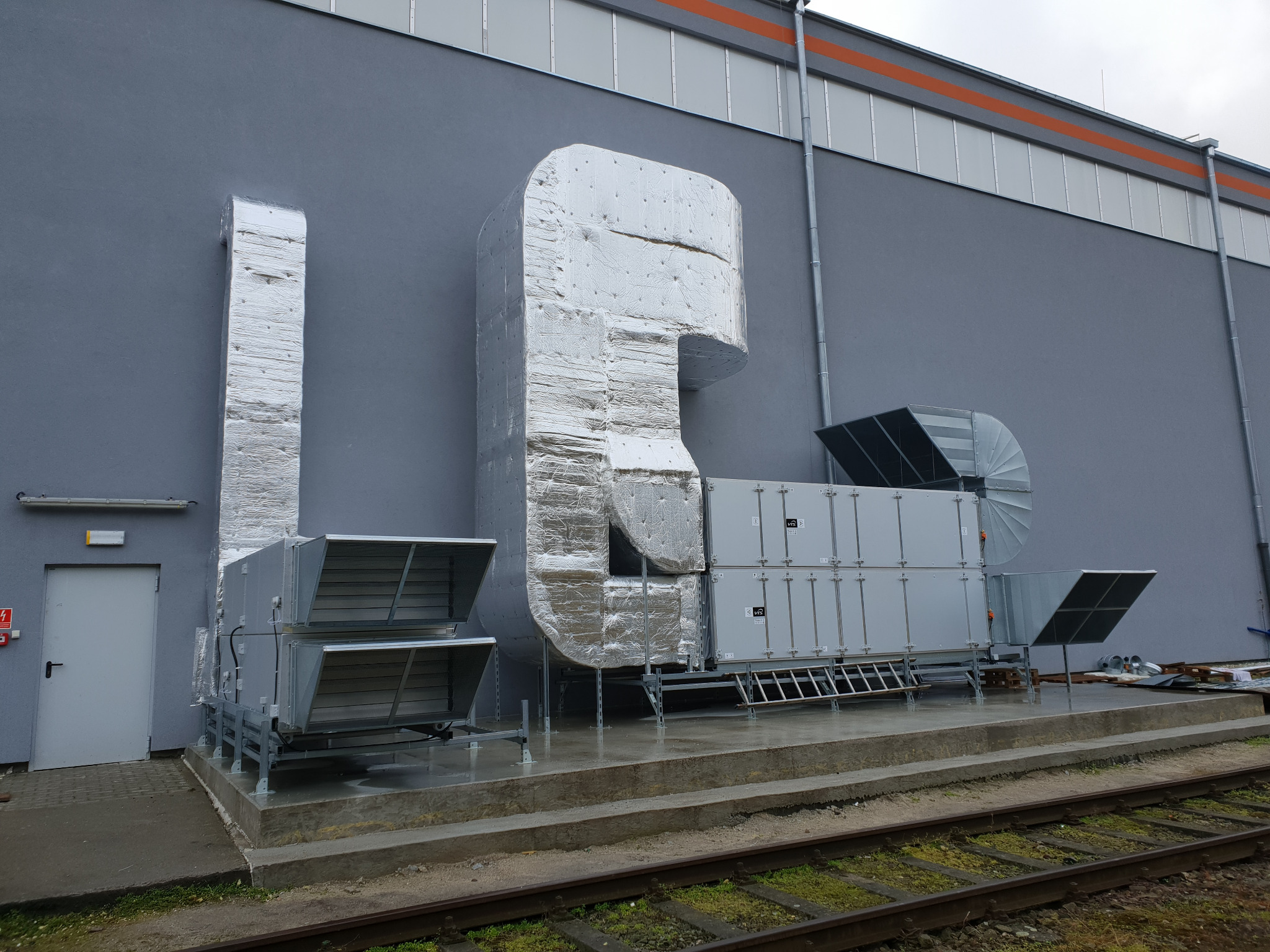 Zewnętrzna instalacja wentylacyjna budynku komercyjnego z izolowanymi kanałami, widoczna szara fasada i fragment torów kolejowych