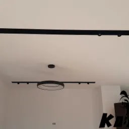 Nowoczesne oświetlenie sufitowe z czarnymi listwami i okrągłą lampą wiszącą w minimalistycznym wnętrzu z oknem po lewej i dekoracyjnym napisem na ścianie po prawej.