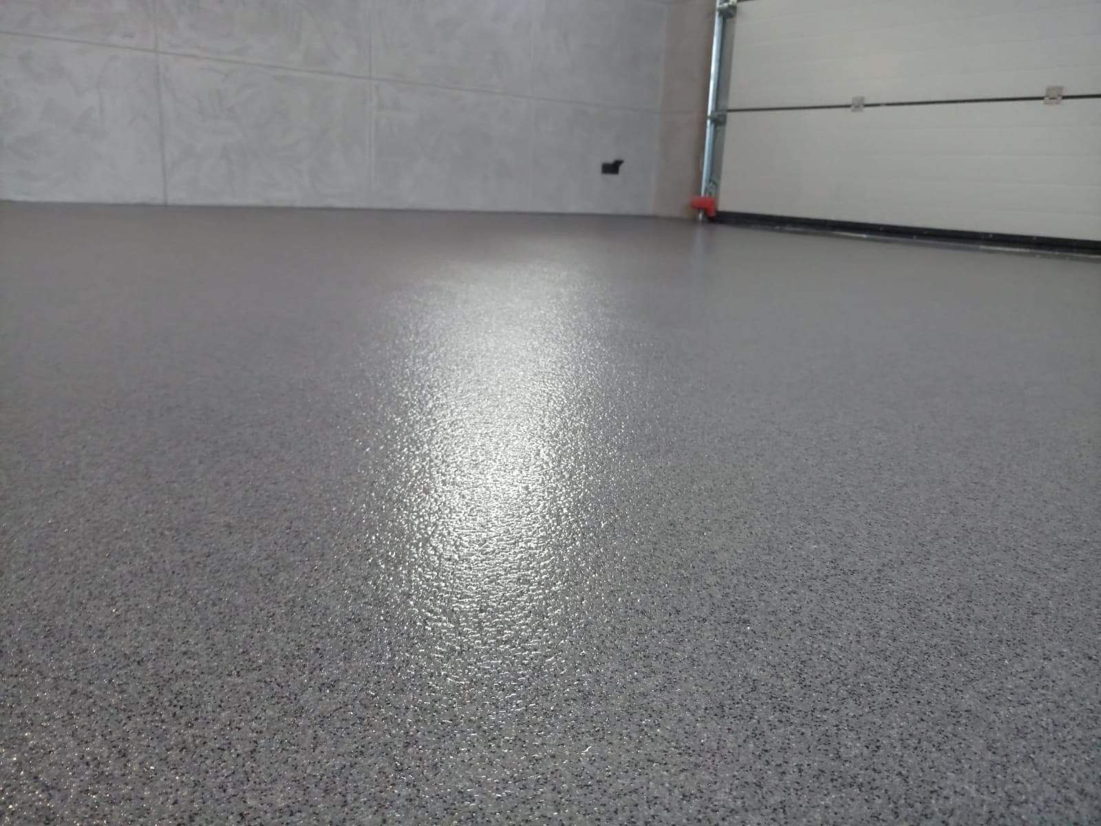 Błyszcząca, szara posadzka żywiczna z delikatnym wzorem w garażu, widoczna brama garażowa z elementami czerwieni.