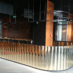 Nowoczesna, złota balustrada z pionowych paneli, otaczająca przestrzeń, z czarną metalową konstrukcją nad nią i pomarańczowo-brązowymi ścianami w tle.