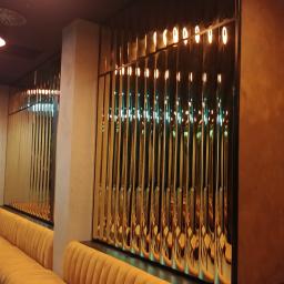 Lasertom Grzegorz Tomaszewski - Dekoracyjna ścianka działowa z pionowych, metalowych listew z oświetleniem, oddzielająca przestrzeń z żółtą, pluszową sofą.