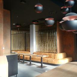 Lasertom Grzegorz Tomaszewski - Wnętrze kawiarni lub restauracji z rzędami złotych, pionowych listew, miękkimi, pomarańczowymi sofami wzdłuż ścian i nowoczesnymi, czarnymi lampami wiszącymi z różowym wnętrzem.