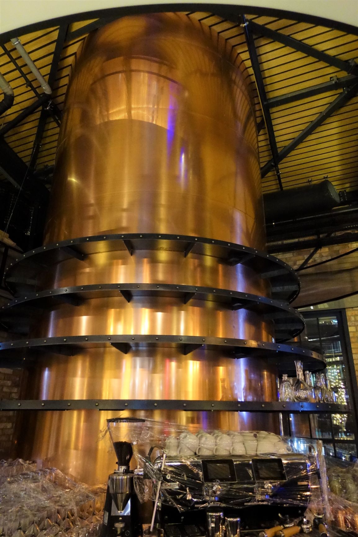 Miedziana kolumna z metalowymi obręczami, prawdopodobnie element dekoracyjny lub instalacja przemysłowa, widoczna w lokalu gastronomicznym w Warszawie, z ekspresami do kawy i szklankami częściowo...