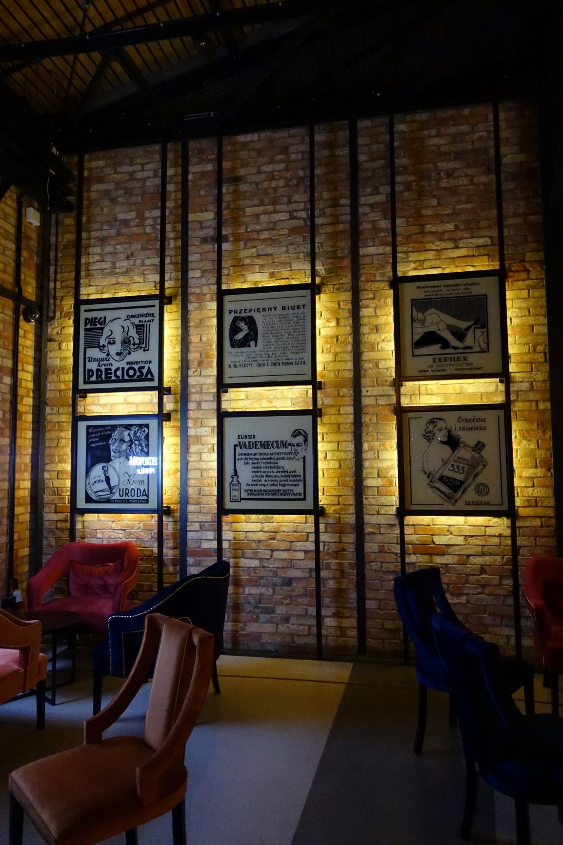 Ściana z cegły z metalową konstrukcją, na której wiszą oprawione w ramy stare plakaty reklamowe, oświetlone od tyłu, w dolnej części widoczne krzesła w różnych kolorach.